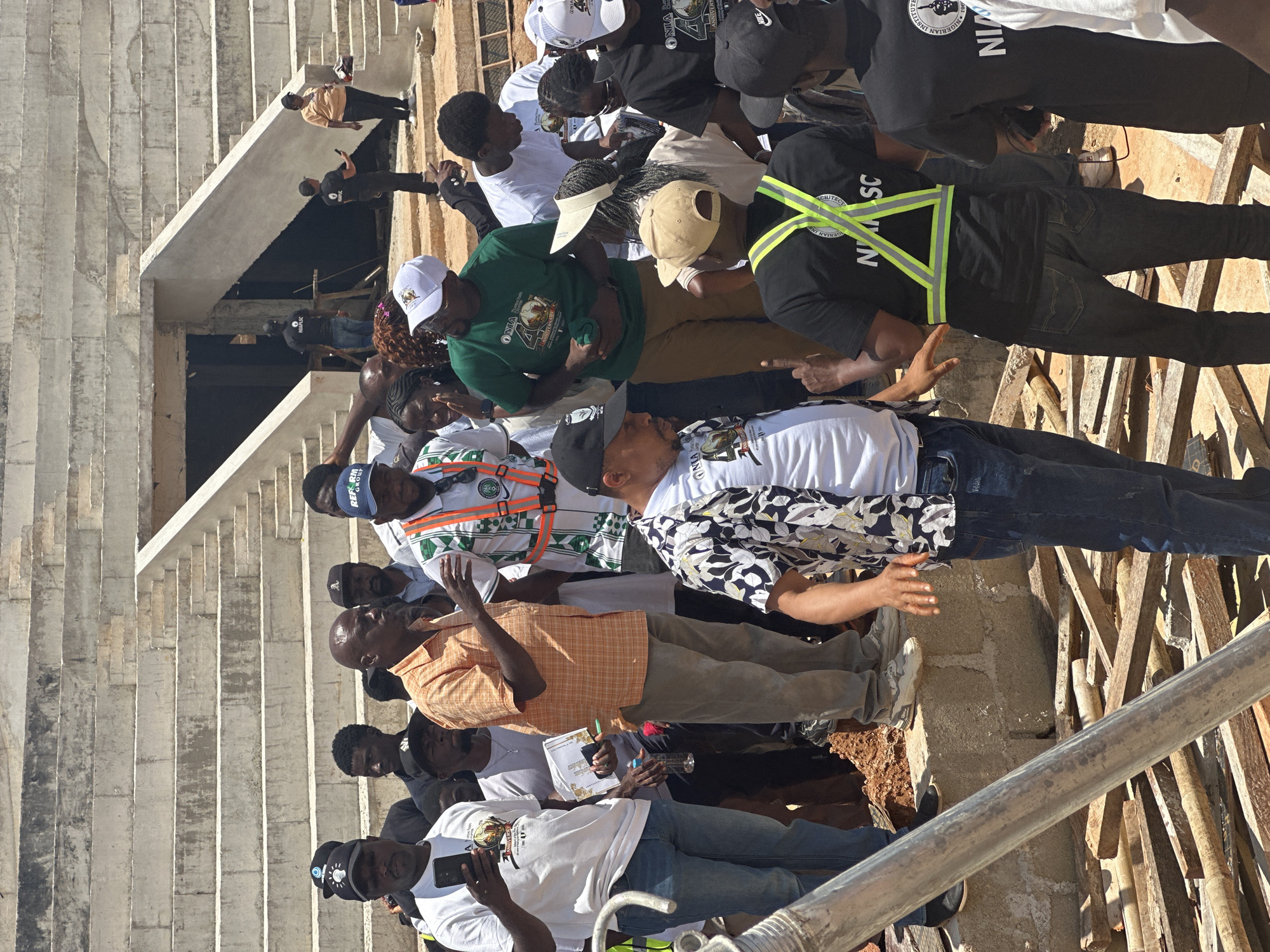 In Jos, Nigeria, a group of individuals, some in construction attire, gather at an unfinished concrete stadium. The stadium seating, visible in the background, is partially constructed and appears weathered. A central figure, wearing an orange checkered shirt and grey trousers, gestures while addressing others. Several men wear t-shirts with various logos, including one that reads "NIAASC" and another with "REFORM GROUP". Some individuals hold phones, suggesting documentation or communication. The ground is uneven, with piles of wood scattered around, indicating ongoing construction work. The atmosphere appears to be one of inspection or discussion related to the stadium project.