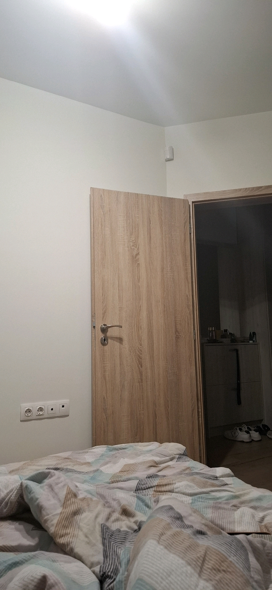 This image depicts a bedroom interior with a bed in the foreground covered with a patterned duvet. To the right, a wooden door is ajar, revealing a glimpse of another room. On the wall to the left of the door, there are electrical outlets. The scene is likely from a residential apartment in Sofia, Bulgaria.