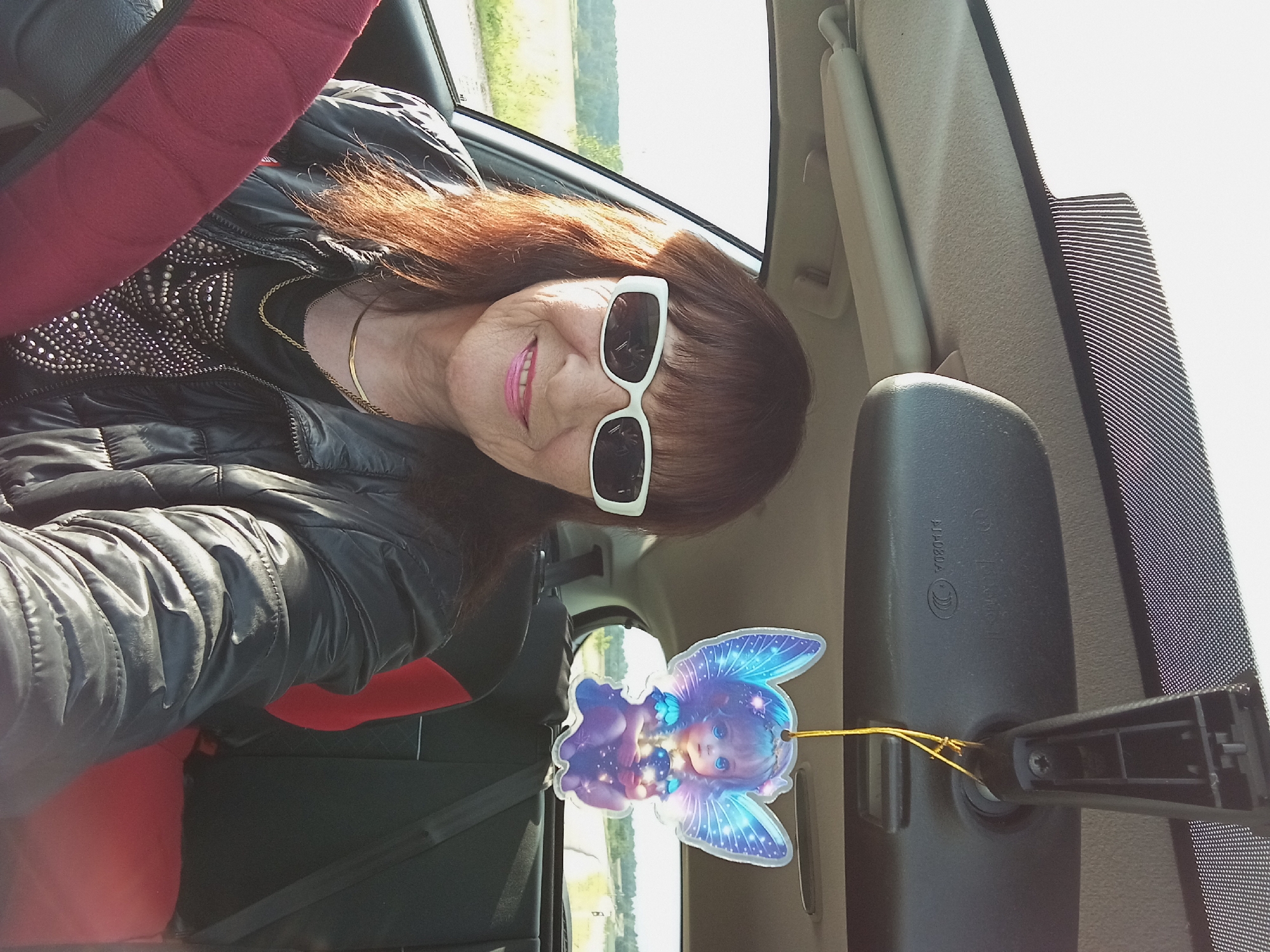 An elderly Caucasian woman with brown hair, sunglasses, and a black jacket is sitting in the driver's seat of a car. She is smiling and looking at the camera. A fairy-shaped air freshener hangs from the rearview mirror. The car's interior is visible, as are the passenger seat and the road outside through the windows. The setting appears to be a rural or suburban area during the day, with a bright sky and trees visible in the distance. The image is a selfie taken from inside the car.