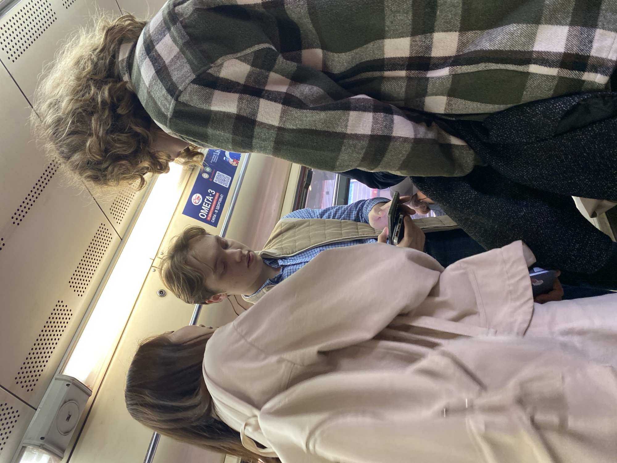 The photo shows a group of people standing in what appears to be a subway car. The individual closest to the camera is wearing a light pink coat. Behind them, a person is looking at a phone, dressed in a blue plaid shirt and a tan quilted vest. Above them, someone is wearing a dark green and white plaid jacket. The environment suggests the interior of a public transit vehicle, with advertisements and metal panels visible.
