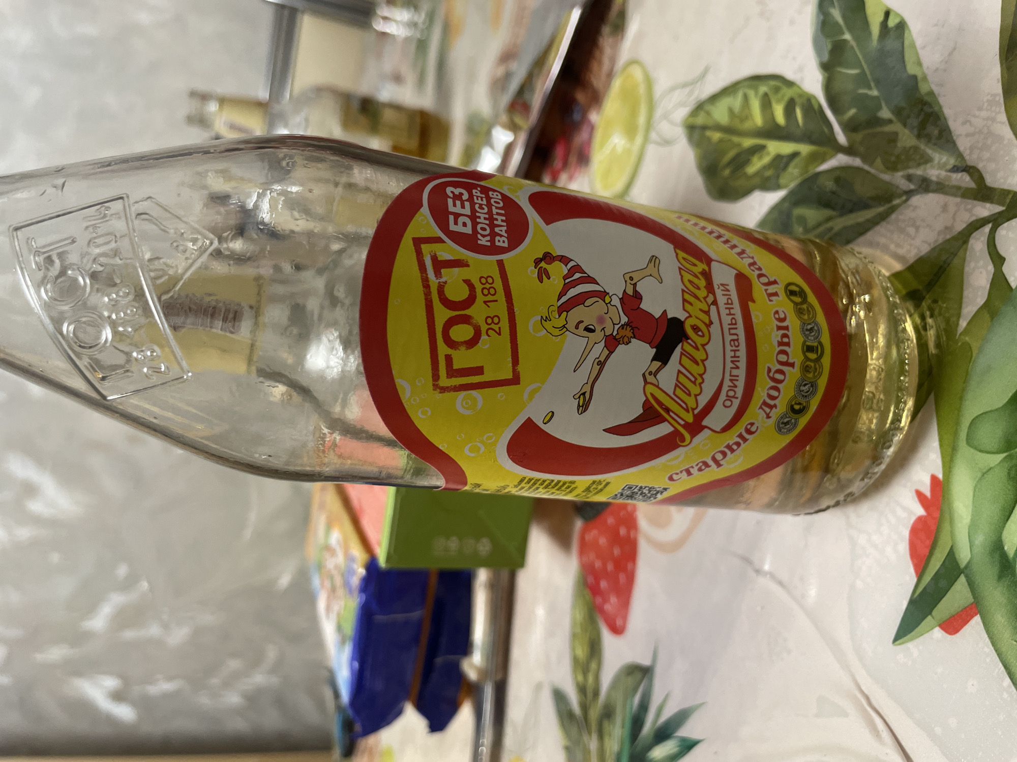 A clear glass bottle of lemonade with a yellow label featuring a cartoon character and Russian text is positioned on a patterned tablecloth. The label has red and yellow elements and reads "ГОСТ 28 188" and "БЕЗ КОНСЕР-ВАНТОВ". Below this, in a wavy font, is "Лимонад" and further text reads "оригинальный старые добрые традиции". The bottle is partially filled with a pale yellow liquid and shows some condensation. In the background, out of focus, are other items, including what appears to be food packaging and another bottle. The overall setting suggests an indoor domestic environment. There is no information in the image to suggest a location in the city of Laishevo, Russia.