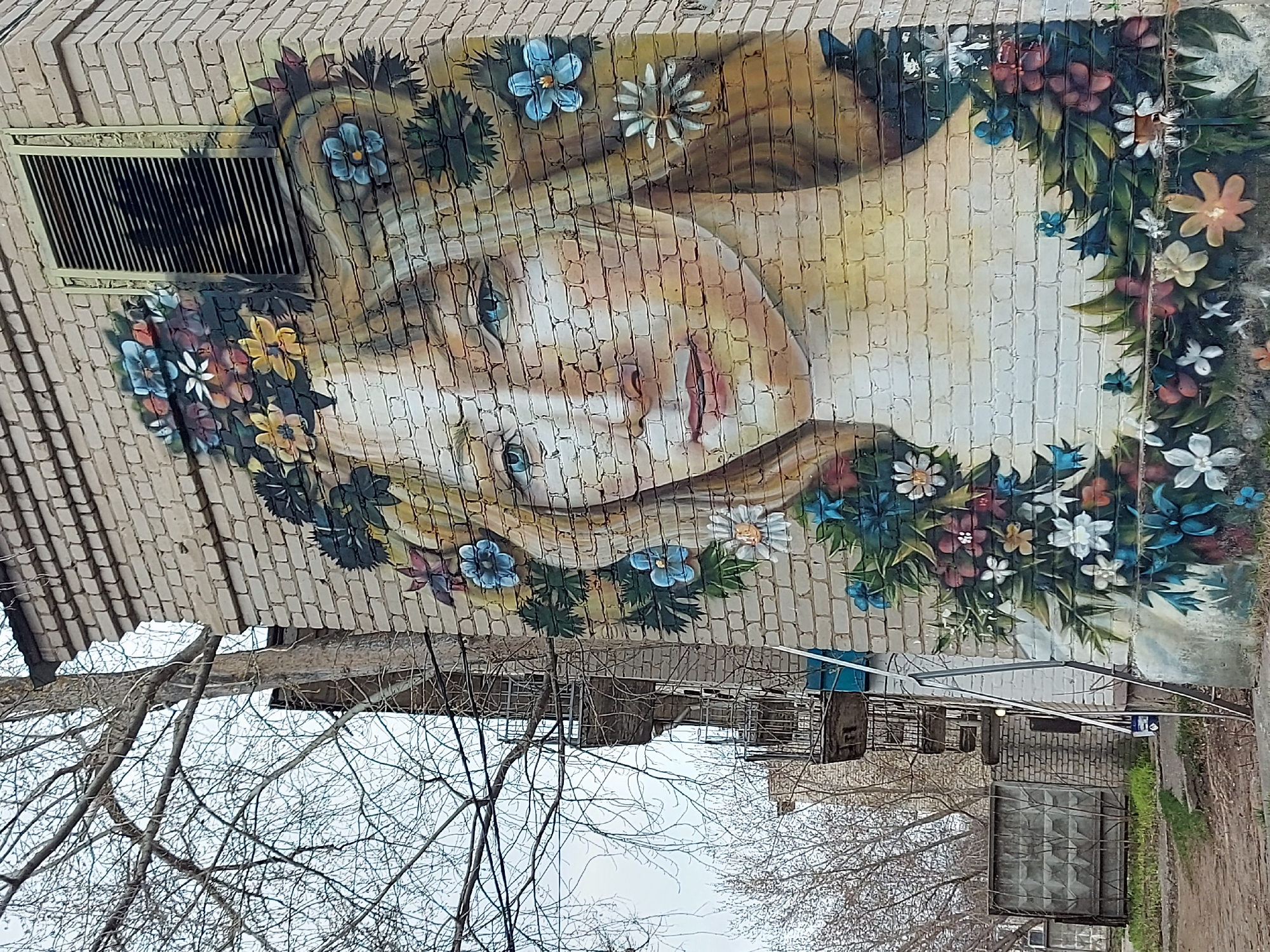 A brick wall in Chaikovsky, Russia, features a mural of a woman with blonde hair adorned with flowers. The woman's face is rendered in a pale palette, and her eyes are blue. A wreath of flowers encircles her head, with various colors and species depicted. An open vent is visible on the upper left corner of the mural. Below the mural, bare tree branches are visible against a muted sky. In the foreground, there is a visible building and its cage-like door.