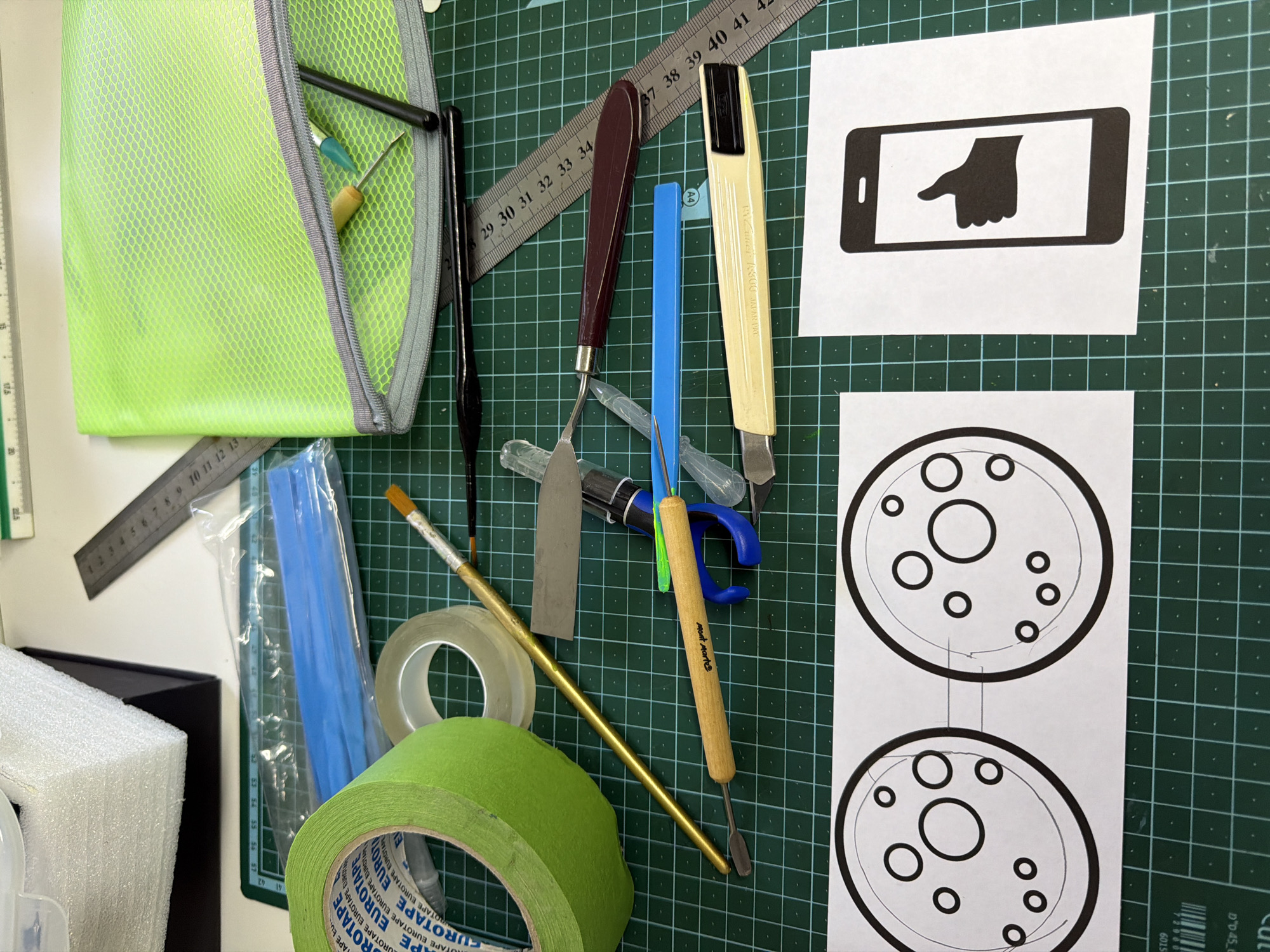 A top-down view reveals an assortment of craft supplies and tools arranged on a green cutting mat. A neon green mesh bag sits on the left, partially obscuring a metal ruler. Various tools are scattered across the mat, including sculpting tools with wooden handles, a palette knife, brushes, and a utility knife with a beige handle. Rolls of transparent and green masking tape are also present. To the right, two pieces of white paper display printed graphics: one depicting a hand pointing at a phone screen and the other showing two circular diagrams with concentric circles. The cutting mat features a grid pattern and appears to be on a table surface, with a white surface and what appears to be a black box partially visible on the left edge of the frame.
This image does not contain information about the city of Even Yehuda, Israel.