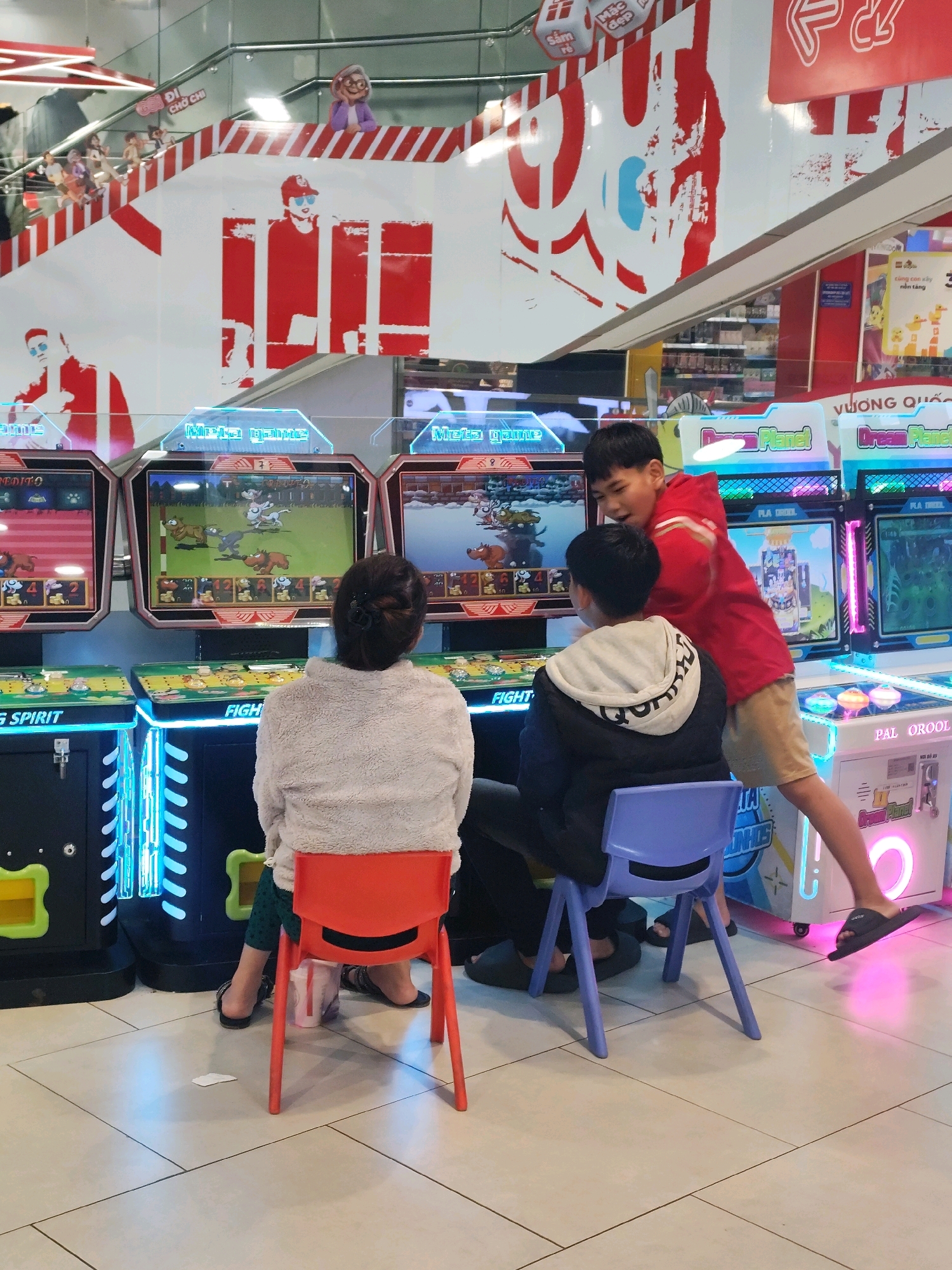 This is a medium shot, eye-level interior photograph taken at an arcade in Da Lat, Vietnam. In the foreground, three people are engaged with arcade games. A woman sits in a red plastic chair facing away from the camera, looking at a game screen. In front of her, two boys stand. One boy, wearing a red hooded jacket, is leaning forward, his arm extended towards the game. The other boy, wearing a black hooded jacket, is seated on a purple plastic chair facing the games. The arcade machines have brightly lit screens displaying animated characters, and the floor is tiled. In the background, a multi-tiered staircase with red and white patterned sides rises upwards, adorned with large red and white graphic designs. Various arcade machines and displays are visible throughout the space. The overall atmosphere is one of a busy entertainment venue.