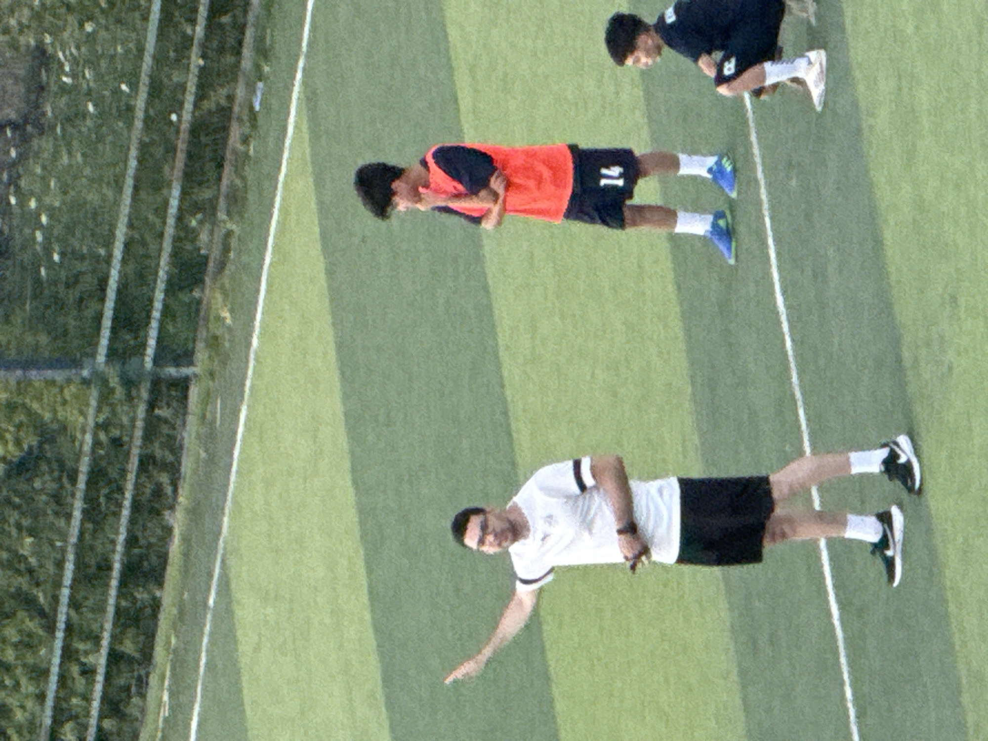 The image depicts an outdoor soccer field with artificial turf. Three individuals are visible on the field. On the left, a man wearing a white t-shirt with black and white striped sleeves and black shorts is standing and gesturing with his left arm. He appears to be a coach or instructor. In the center, a young person in an orange vest over a dark shirt and dark shorts with the number "14" is standing with arms crossed and chin resting on their hand, appearing to be listening or observing. On the right, another young person is partially visible, crouching or sitting on the grass in a dark blue jersey and dark shorts. The background shows a fence and dense green foliage. The scene takes place on a soccer field in the city of Tbilisi, Georgia.