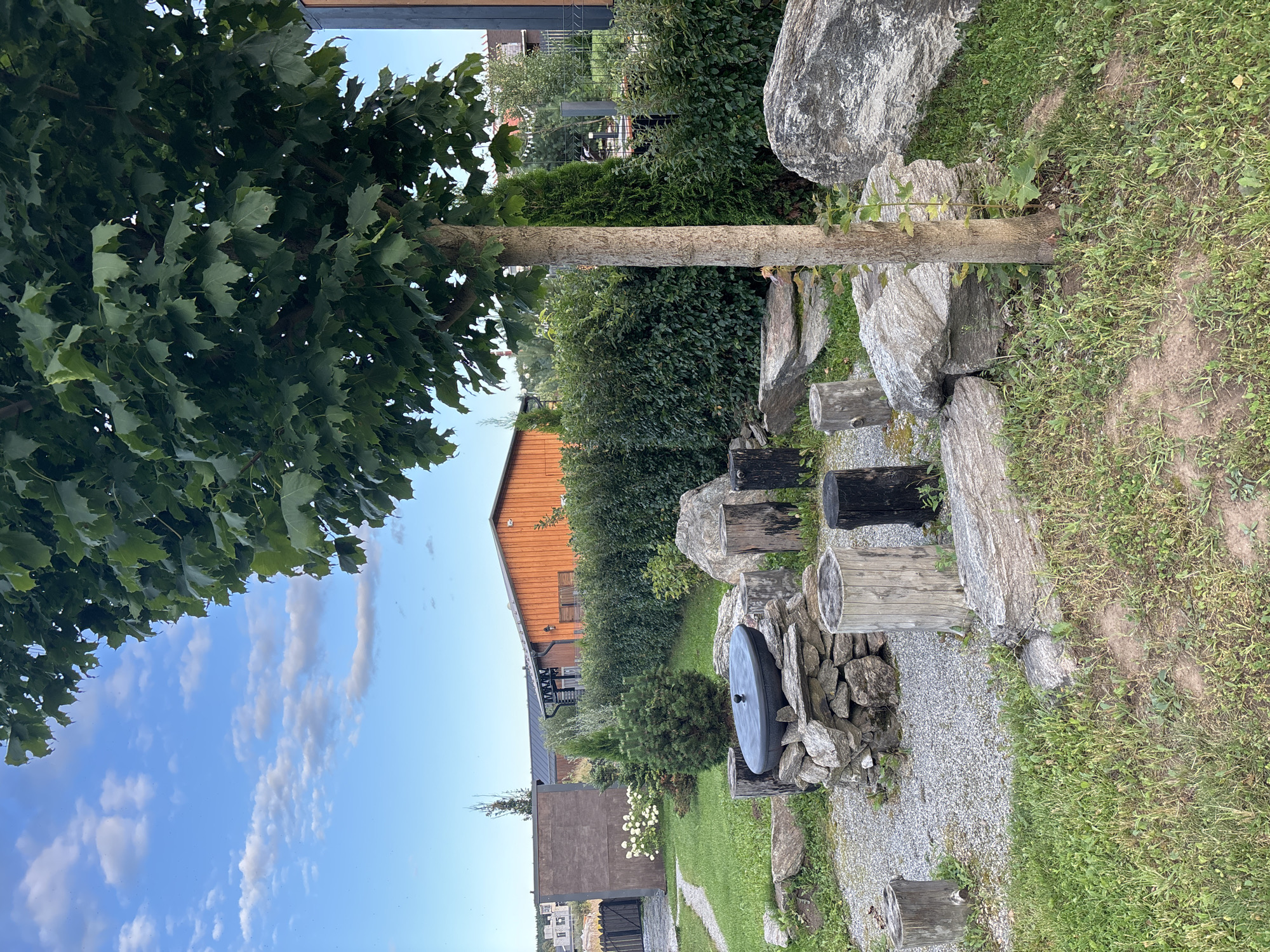 The image depicts an outdoor scene in Zvenigorod, Russia. In the foreground, a stone fire pit is arranged with a large black pot on top, surrounded by several tree stumps and large rocks. A young tree stands to the right of the fire pit. Behind the fire pit, a well-maintained hedge forms a backdrop, with an orange-colored building visible beyond it. The ground is covered with green grass and gravel pathways. The sky is blue with scattered clouds. The scene appears to be a backyard or garden area.