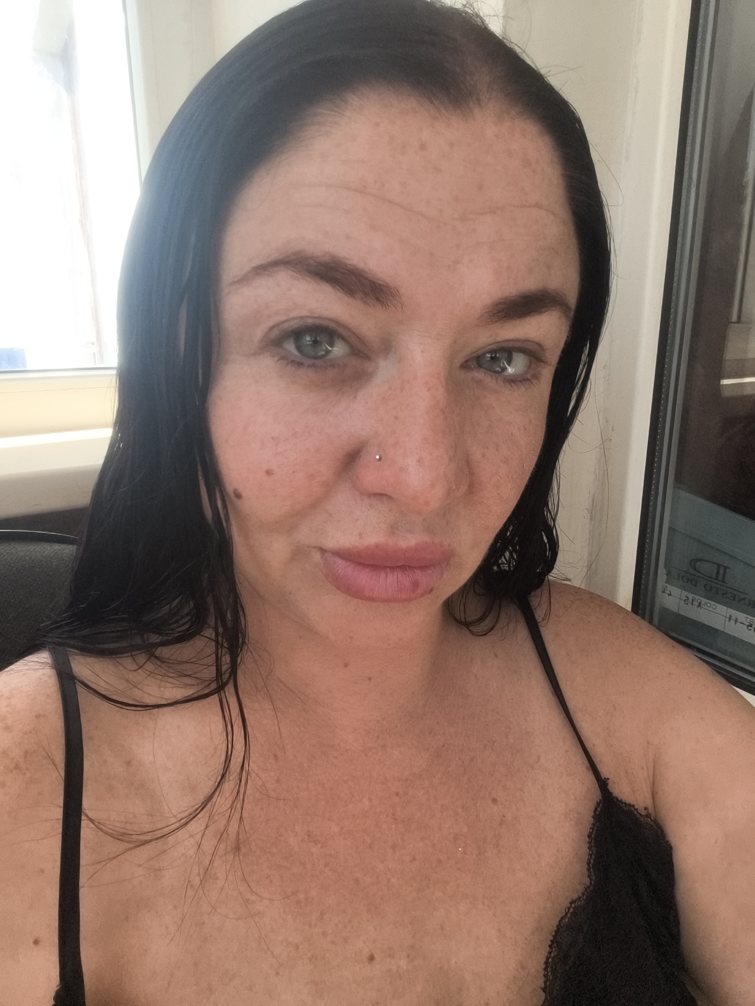 A woman with dark hair and fair skin with freckles is shown from the chest up, looking directly at the camera. She is wearing a black camisole with lace trim. She has a nose piercing with a small silver stud. The background appears to be an indoor setting with a window on the left side of the image and a window with a reflection on the right. The lighting is bright, suggesting it is daytime. No specific landmarks of Lisakovsk, Kazakhstan are visible.