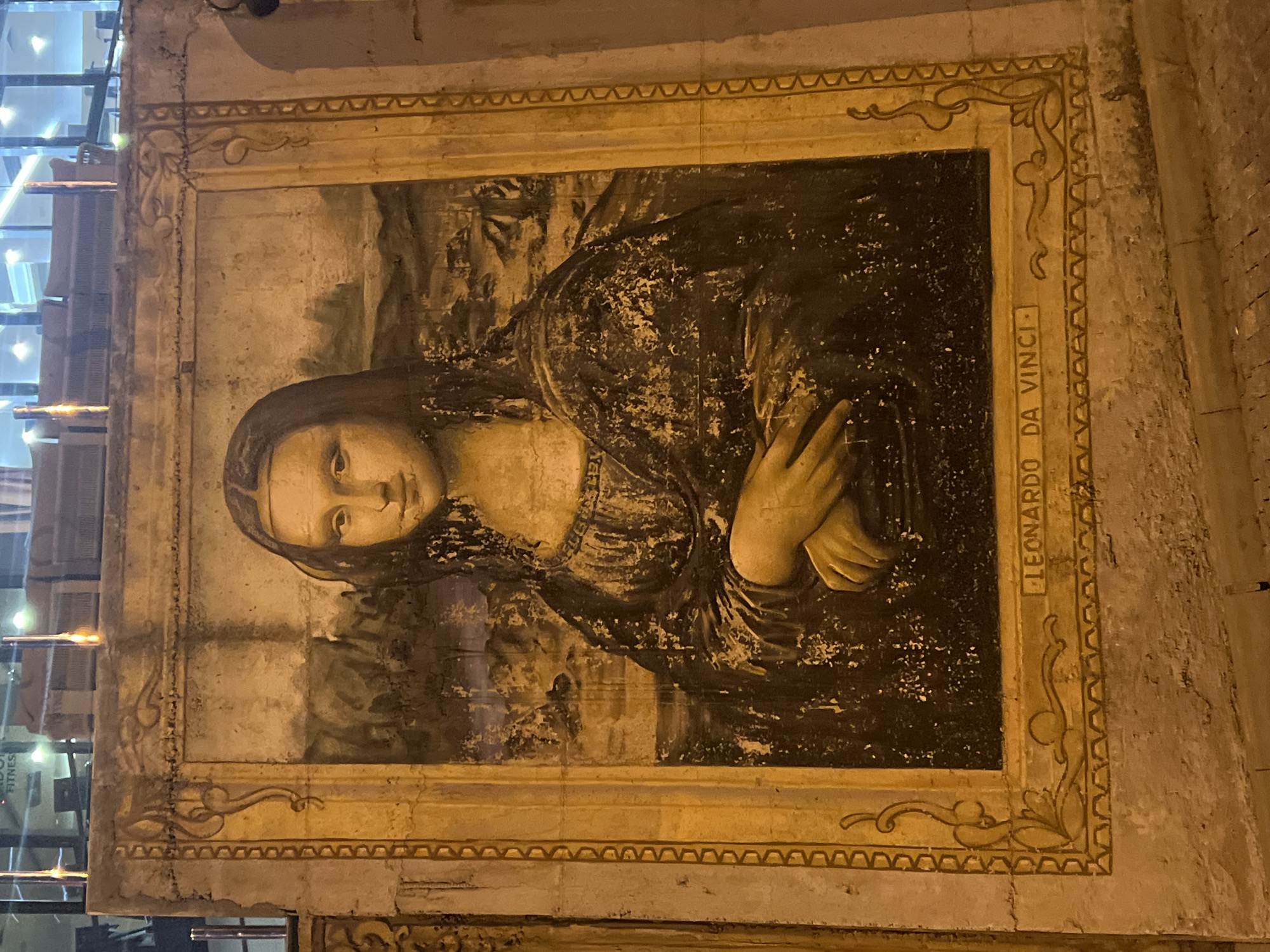 This image depicts a mural of the Mona Lisa painted on a textured, weathered wall in Odesa, Ukraine. The mural is framed by a decorative border that appears to be painted directly onto the wall. The name "LEONARDO DA VINCI" is inscribed in capital letters within the lower border of the frame. The painting itself shows signs of wear and tear, with visible cracks and discoloration on the wall surface, suggesting it is an outdoor artwork exposed to the elements. In the background, above the mural, the facade of a modern building with large glass windows and internal lighting is visible, indicating an urban setting. The lighting in the scene suggests it is nighttime or dusk.