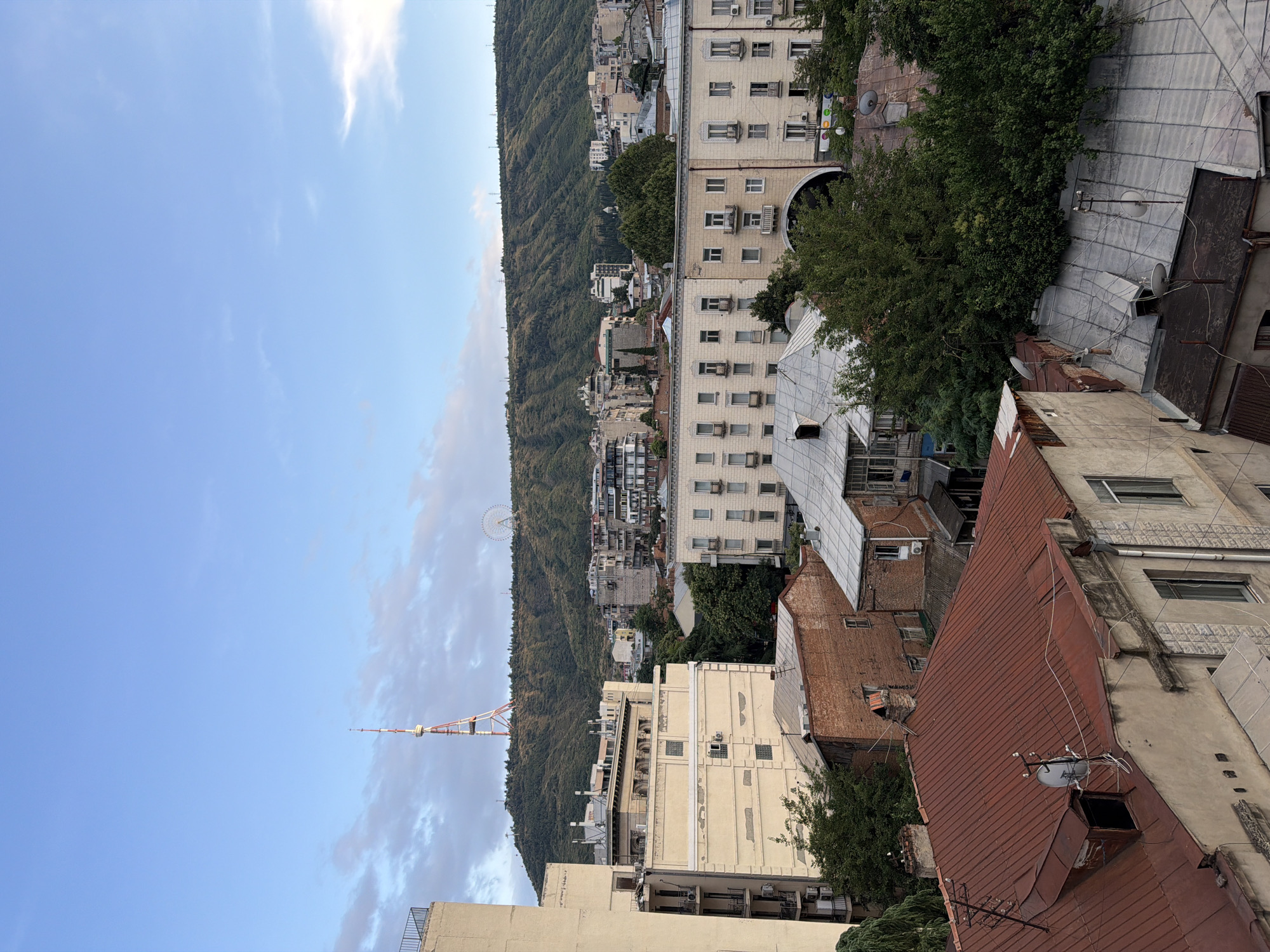 This is an elevated, eye-level, medium shot of an urban landscape in Tbilisi, Georgia. In the background, a large, green, tree-covered hill rises, topped with a prominent radio tower and a Ferris wheel. The middle ground is filled with a dense cluster of multi-story buildings, varying in architectural style and color, including cream-colored apartment blocks with balconies and arched entrances, and darker, more modern structures. In the foreground, several older buildings with rust-colored and grey corrugated metal roofs are visible. Trees are interspersed throughout the scene, with lush green foliage contributing to the overall impression of a city nestled within a natural environment. The sky is a clear blue with scattered white clouds, suggesting fair weather. There are no people or animals visible in the image.