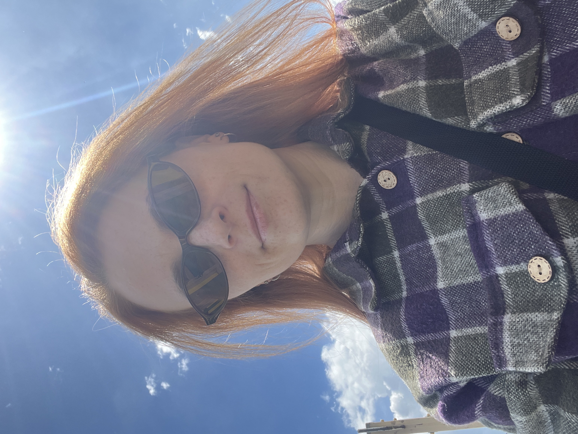 A selfie, taken from a low angle looking up, shows a person with red hair wearing sunglasses and a purple and gray plaid shirt with wooden buttons. A black strap crosses their body. The background features a blue sky with some clouds and a bright sun. Lower right of the frame are some wires of unspecified type running between utility poles. The location is in Igra, Russia.