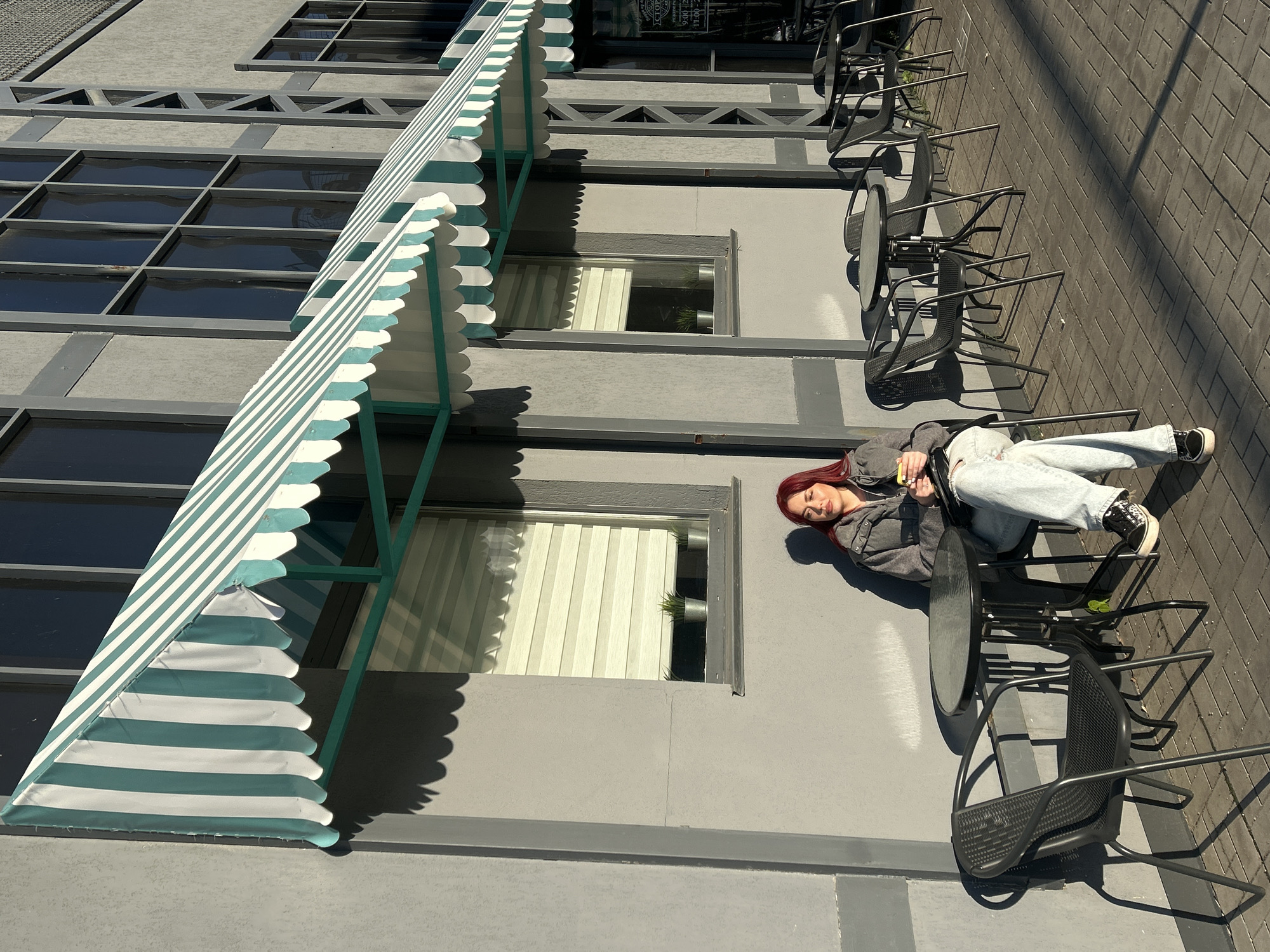 A young woman with long red hair lies on her side on a gray paved surface next to an outdoor cafe setup. She wears a gray hooded jacket, light blue wide-leg jeans, and black and white sneakers. Her eyes are closed or looking downwards, and she holds a yellow object, possibly a phone, in her left hand.
The scene depicts an outdoor area in front of a gray building, likely a commercial establishment in Tolyatti, Russia. The building features numerous large rectangular windows, some of which show reflections, while others reveal interior details like white vertical blinds and small potted plants. A prominent teal and white striped awning with a scalloped bottom edge extends from the building's facade, casting distinct shadows.
Several black metal outdoor tables and chairs are arranged on the dark gray brick-patterned pavement. Strong sunlight indicates a bright daytime setting, with long shadows cast by the furniture, awning, and the woman. Faint text and what appear to be business hours ("9:00 - 20:00" and "11:00 - 21:00") are visible on a sign or window behind the awning, suggesting the establishment is open. The overall atmosphere is relaxed and sunny.