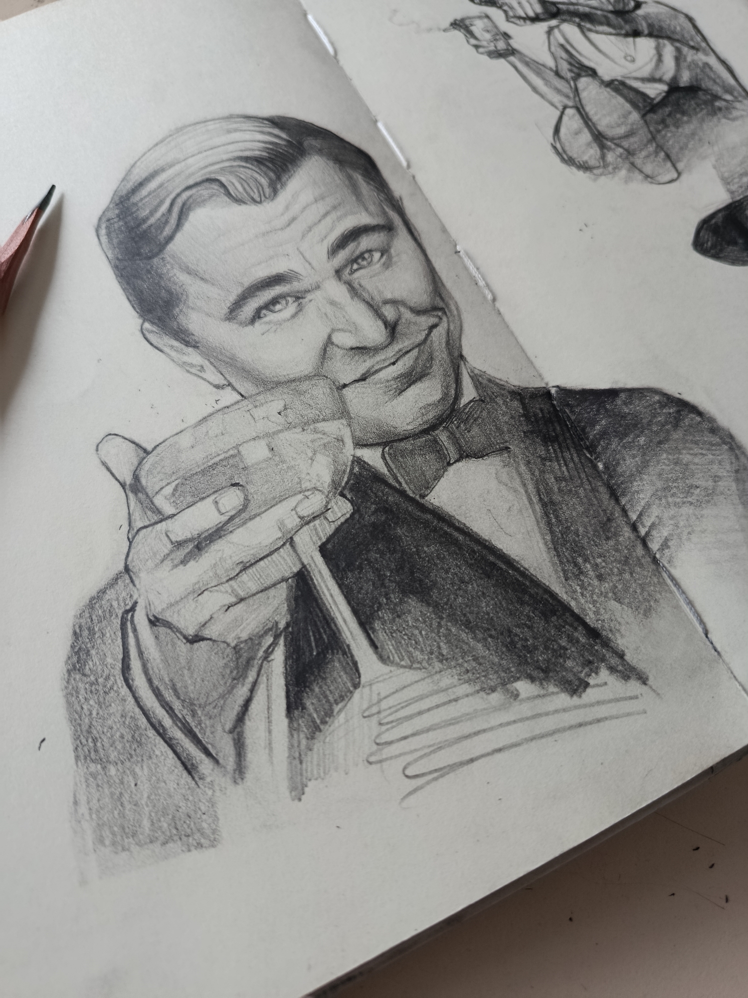 A close-up photograph captures a pencil drawing in a sketchpad, primarily featuring a man resembling actor Leonardo DiCaprio in his role as Jay Gatsby. The man is depicted from the chest up, wearing a formal suit with a prominent bow tie. He has slicked-back hair and a slight, coy smirk, looking upwards and to his right. His right hand holds a stemmed cocktail glass, which appears to contain liquid, possibly with ice, as if raising a toast or offering a drink. The drawing is rendered in varying shades of graphite, showcasing detailed shading for his hair, facial features, suit, and the glass.
To the left of the main drawing, the sharpened tip of a wooden pencil is visible, resting on the sketchpad, suggesting the medium used for the artwork. In the upper right corner of the sketchpad, a smaller, less detailed pencil sketch of another person is partially visible. This figure appears to be sitting or crouching, holding an object that might be a cigarette, with faint wisps of smoke drawn above it.
The drawings are on off-white or light cream paper, which exhibits some faint graphite smudges and specks, characteristic of a working sketchpad. The binding of the sketchpad is visible along the right edge of the main drawing. The background is a plain, light surface, likely a desk or table. There is no visible text in the image.