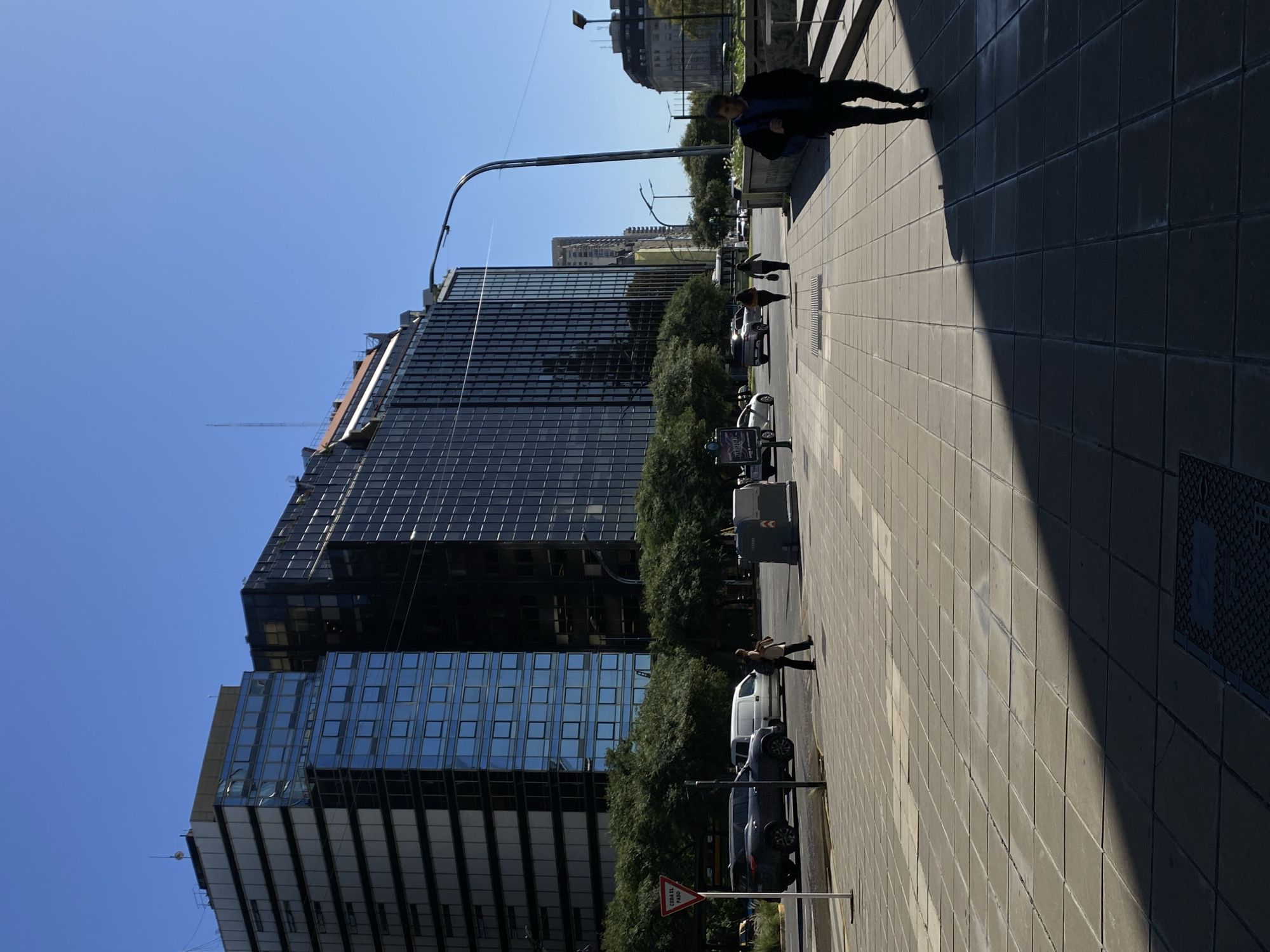 The image captures a sunlit urban street scene in Buenos Aires, Argentina, characterized by modern architecture and daily activity. On the left, towering multi-story buildings with dark, reflective glass facades dominate the skyline, mirroring the bright blue sky. Some scaffolding or construction elements are visible on the upper sections of these structures. A line of green trees runs along the base of the buildings, separating them from the street.
To the right, a wide, paved sidewalk stretches into the distance, populated by several pedestrians. In the foreground, a person in a dark jacket walks away from the viewer. Further down the sidewalk, more people are visible near parked cars. The street itself contains several vehicles, including a white van and a dark SUV, parked alongside the curb. A tall, arching streetlight pole extends over the street from the right side of the frame.
The time of day appears to be midday or early afternoon, with a clear blue sky overhead and bright sunshine casting strong, distinct shadows. A prominent, sharp shadow diagonally cuts across the right half of the pavement, contrasting with the brightly lit areas. A red triangular traffic sign is visible near the bottom center, bearing the Spanish text "CEDA EL PASO," which means "GIVE WAY." The overall scene depicts a bustling, contemporary city environment.