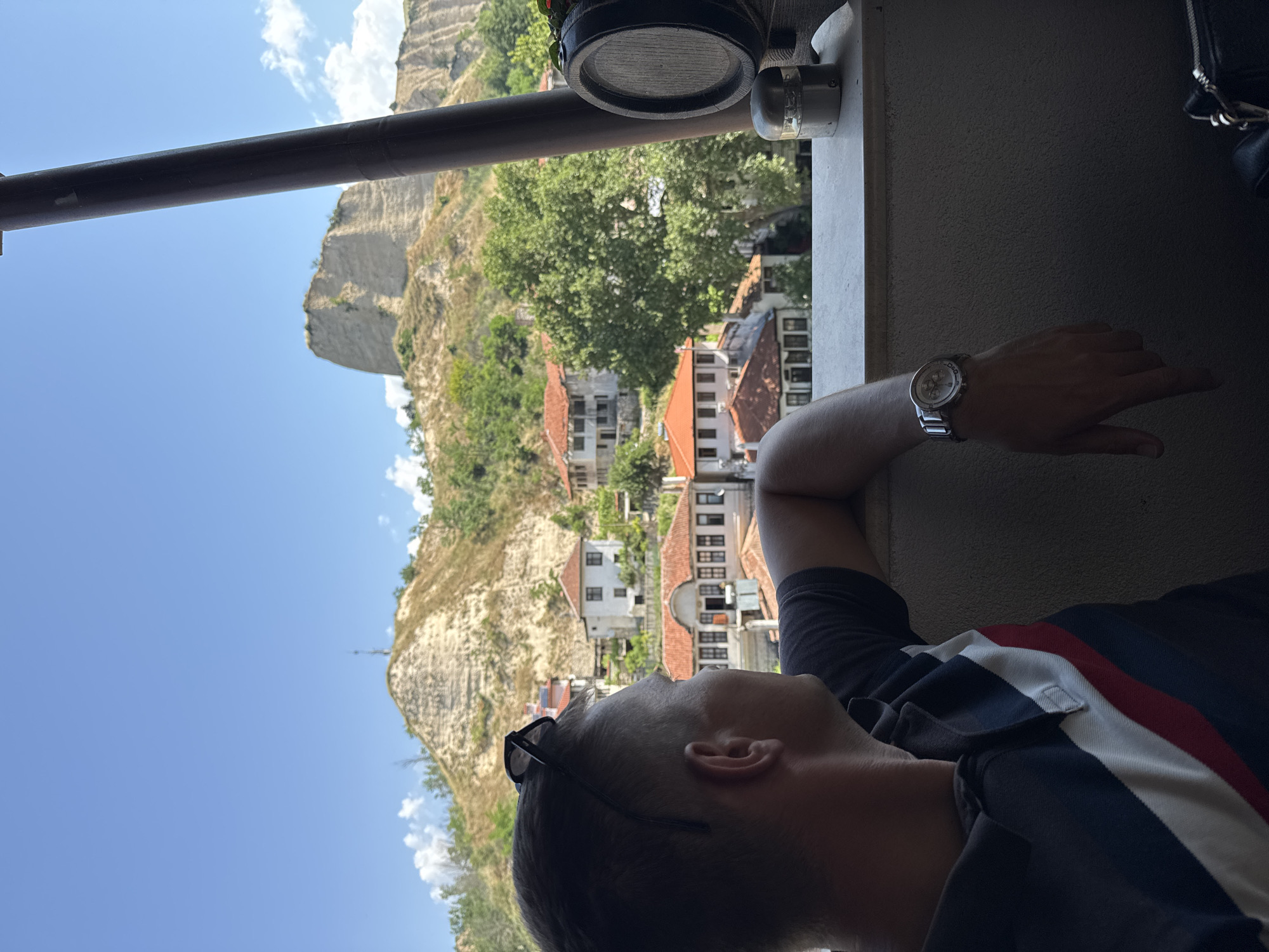 A male individual is observed from a rear-right perspective, gazing towards a panoramic view. He is wearing a dark polo shirt with a vertical white stripe bordered by a red stripe on his left side, and black sunglasses are positioned on his head. A silver-toned wristwatch is visible on his left wrist, which rests on a light-colored concrete parapet. To the far right, a dark cylindrical structural pillar extends vertically. On the parapet, a small wooden decorative barrel with a metal band and a small cylindrical metallic object are present. The middle ground reveals the city of Melnik, Bulgaria, characterized by traditional houses with orange-red tiled roofs, light-colored walls, and numerous windows, interspersed among mature green trees. The background is dominated by prominent, light-colored sandstone formations, consistent with the Melnik Earth Pyramids, which are partially covered with sparse green vegetation, all set against a clear blue sky with scattered white cumulus clouds.