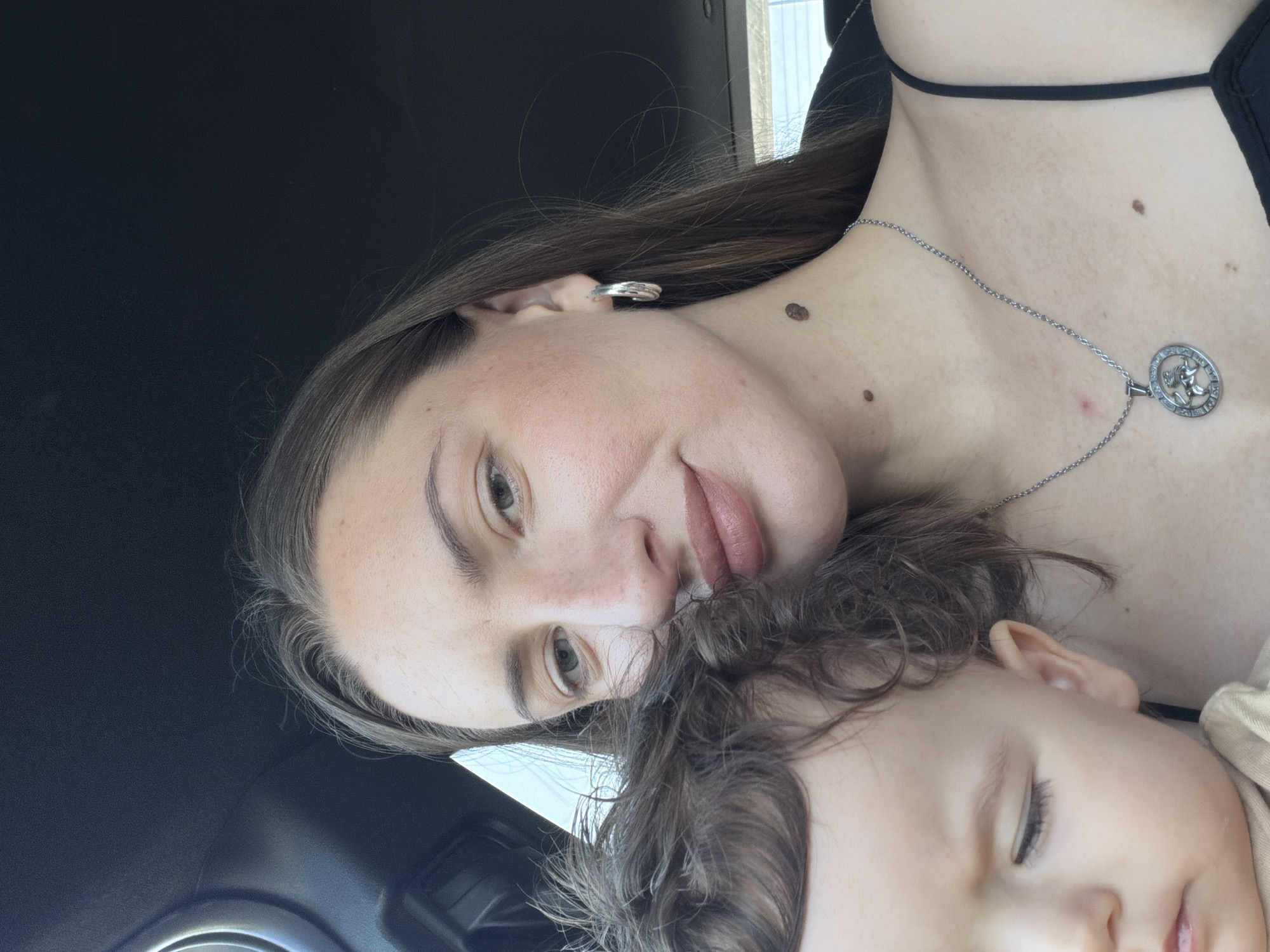 A close-up, selfie-style photograph shows a woman and a baby inside a vehicle. The woman is in the foreground, looking towards the camera with a slight smile. Her long, brown hair is partially obscuring the baby. She is wearing a black spaghetti strap top and a silver pendant necklace. A large mole is visible on her neck. The baby, on the left side of the frame, is turned away from the camera, with its eyes closed and hair curly. The interior of the car, including the headliner, is visible in the background. The photograph does not contain any visual cues to indicate it was taken in Sharm El Sheikh, Egypt.