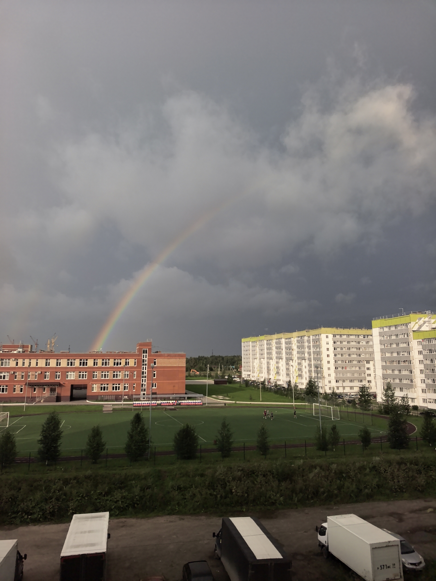 The image shows a residential area in Froli, Russia, with a school and a sports field. A rainbow is visible in the sky, with dark storm clouds in the background. The school building is made of red brick and has many windows. The sports field is green and has a soccer goal. In the foreground, there are several parked trucks. There are some people on the sports field. The buildings in the background are white and tall, with green accents. The overall scene suggests a recent storm that has passed, leaving a rainbow as a sign of clearing skies.