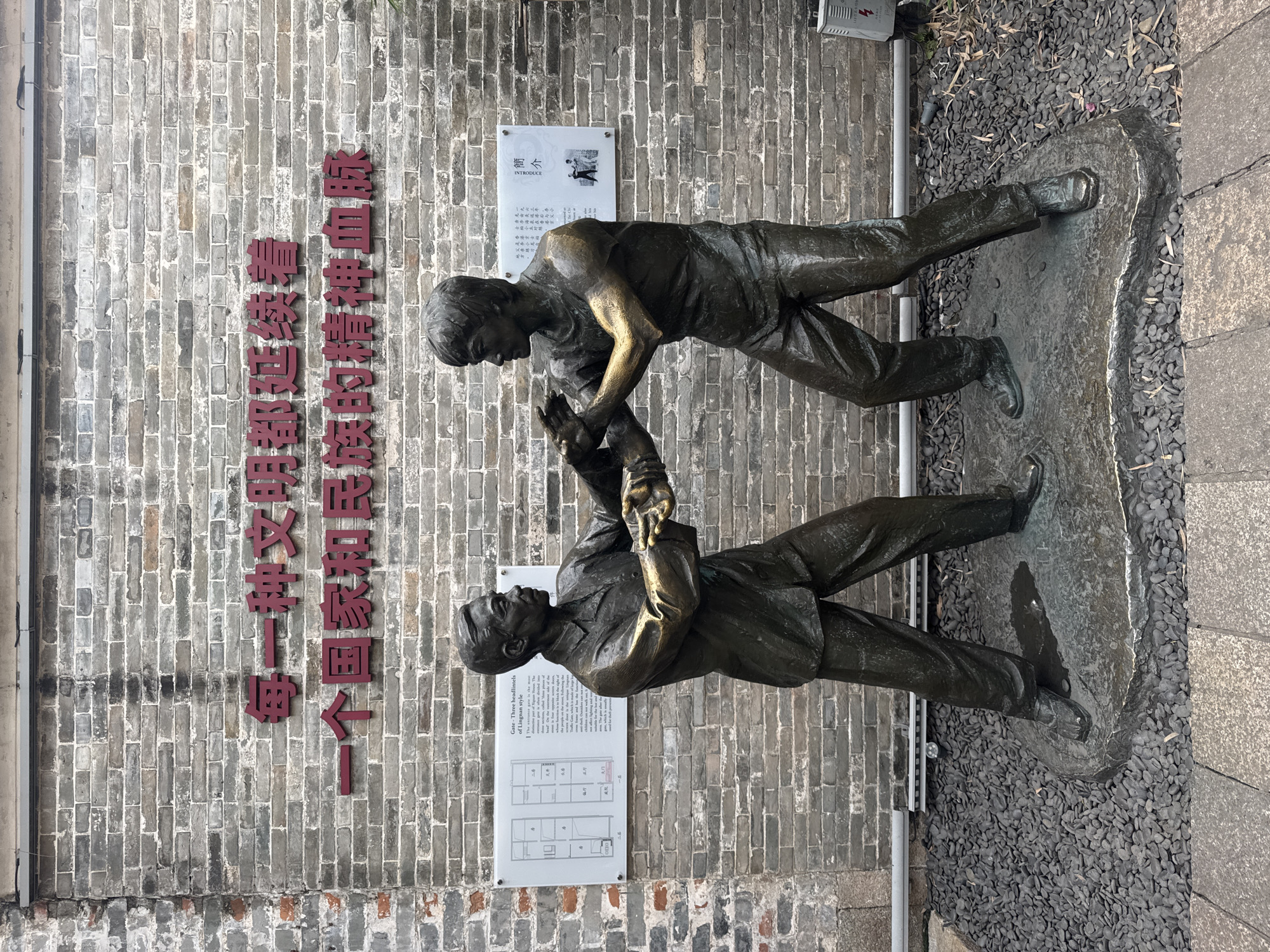 The image shows a bronze statue of two men in front of a brick wall with Chinese characters in the Liwan District of China. One man, dressed in casual clothing, appears to be helping up another man dressed in a suit. Both statues are attached to a stone slab that sits on small, dark rocks next to a stone curb. Two plaques are mounted on the brick wall. The first plaque gives a brief introduction in English and Chinese. The second plaque has Chinese characters and a black and white picture of the statues.