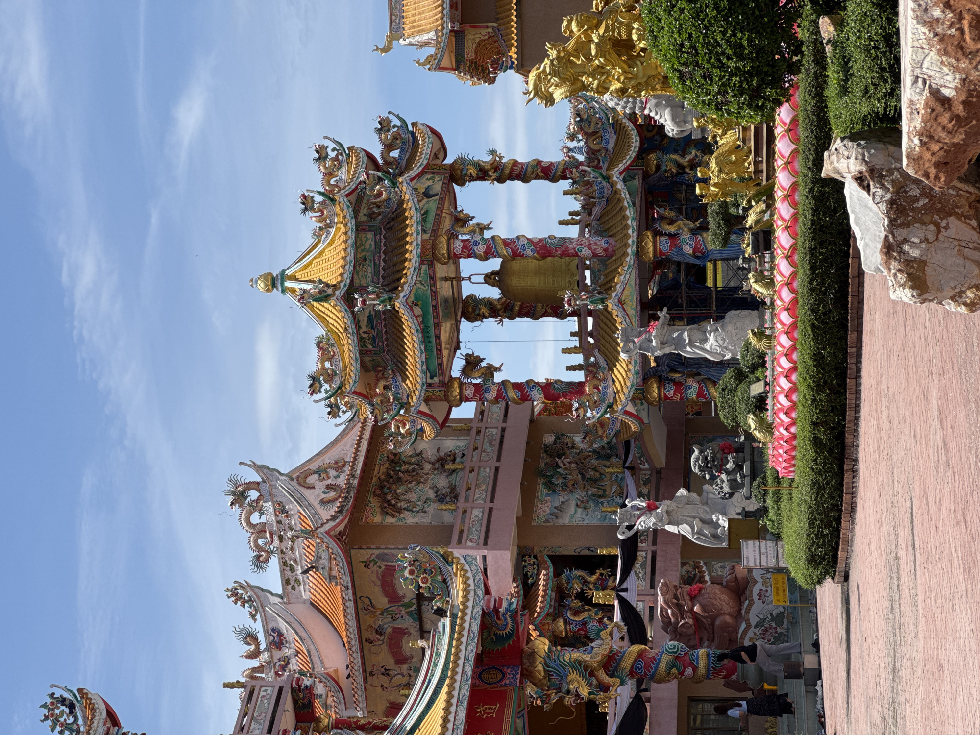 In Ang Sila, Thailand, this image captures a vibrant Chinese temple under a clear blue sky with sparse clouds. The temple's architecture is characterized by ornate, multi-tiered roofs adorned with colorful dragons and intricate carvings. A large, golden bell hangs prominently within a pavilion supported by red and white spiral columns, also decorated with dragon motifs.
In the foreground, a red brick courtyard leads to a manicured garden area featuring low green bushes and a decorative border of pink lotus-like structures. Several stone sculptures are visible, including white figures and guardian lion statues. To the right, a large golden sculpture of mythical creatures stands.
The temple's facade is richly decorated with murals depicting figures and scenes, further enhanced by dragon sculptures on pillars and along the rooflines. In the middle ground, several individuals are seen walking on steps near the temple entrance. The overall scene conveys a sense of cultural richness and spiritual significance, bathed in bright daylight.