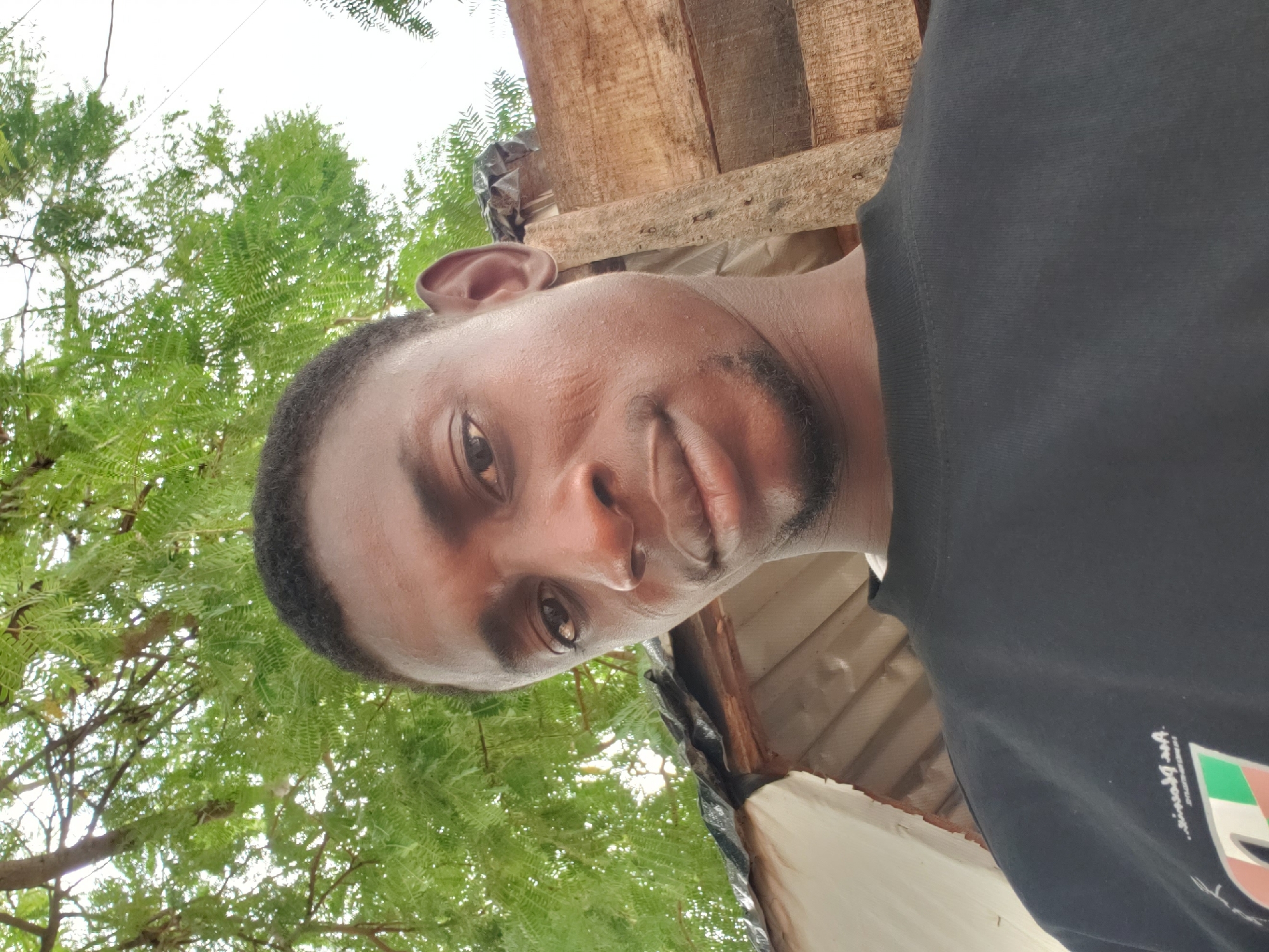 A man with short dark hair and a dark complexion is looking directly at the camera. He has a trimmed beard. He is wearing a dark shirt with a design on the lower right corner. Behind him, there is green foliage suggesting trees. Above him, there is what appears to be the underside of a simple wooden structure, possibly part of a building or shelter. Based on the request, the location is presumed to be Adankolo, Nigeria, but without further information, this is not visible in the image itself. There is no clear evidence of interaction or specific actions happening beyond the man posing for the picture.