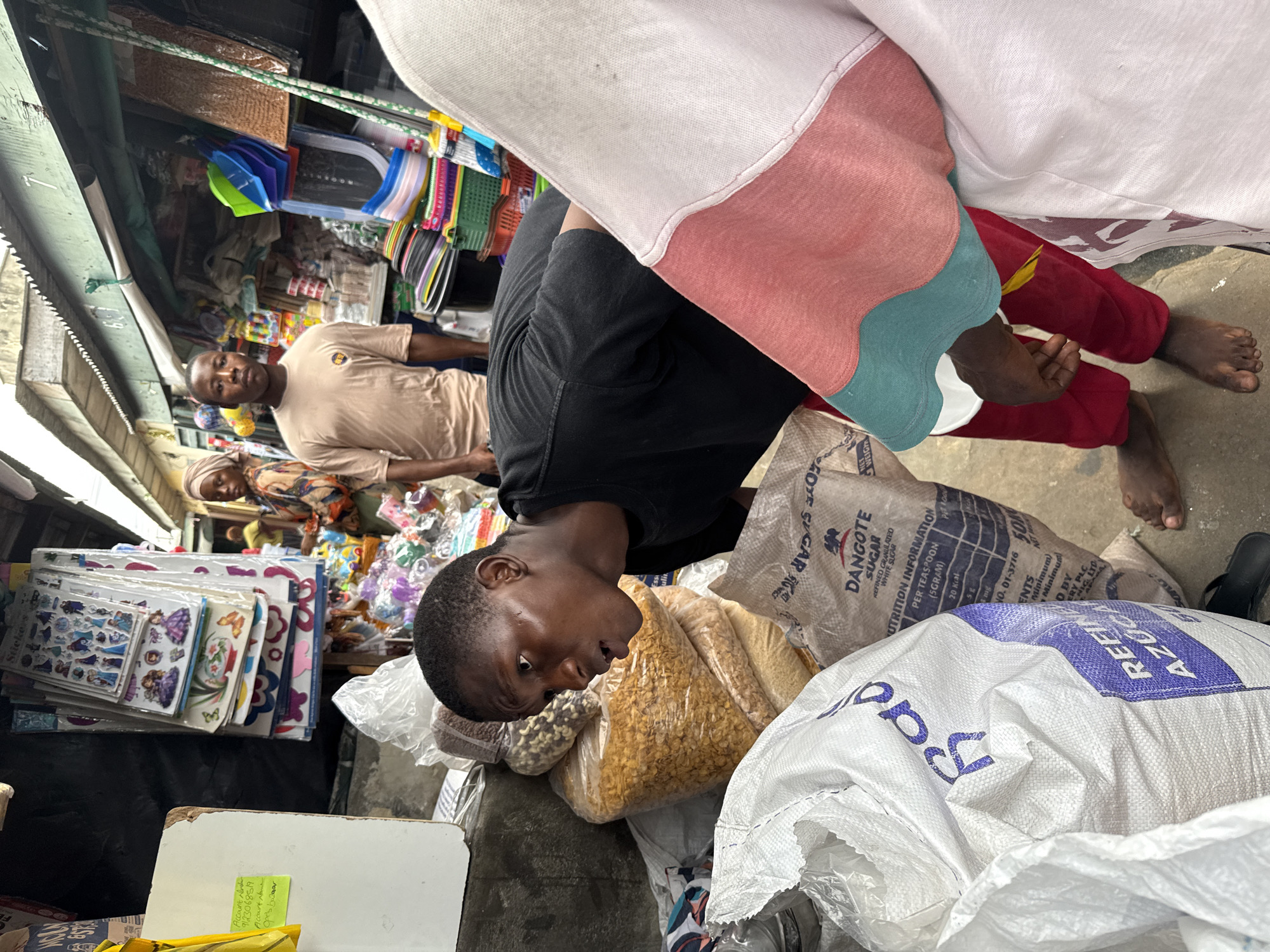 This media file captures a vibrant scene within a market in Lekki, Nigeria, likely during the daytime. The perspective is from above, looking down into a stall or aisle.
In the foreground, a young man wearing a black t-shirt is partially visible, leaning over large sacks of goods. He appears focused on the items in front of him, possibly arranging them. Next to him, another person is seen from the waist down, wearing red trousers and a multi-colored top (white, pink, and teal sections), with bare feet on the ground.
In the midground, a man in a light brown t-shirt stands, looking directly towards the viewer. Behind him, a woman in a patterned top and headscarf is partially visible, also appearing to be part of the market activity.
The setting is a bustling market stall, characterized by an abundance of merchandise. Various plastic items, household goods, and packaged products are displayed on shelves and hanging. Bags of grains or food items are visible near the foreground figures. A stack of decorative stickers, featuring designs like "Frozen" characters and butterflies, is also present.
Visible text on the sacks includes "DANGOTE SUGAR," indicating "REFINED GRANULATED WHITE SUGAR," along with "NUTRITION INFORMATION," listing "PER TEASPOON (5GRAM), 20 Kcal, 5g." Another sack bears the word "AZUCAR" and "Rad." On a white surface, handwritten text for an "Account" with the number "9123068519" and "Account Name: Opels bowaar" is visible on a sticky note. The overall atmosphere suggests a busy commercial exchange in an open or semi-open market environment.