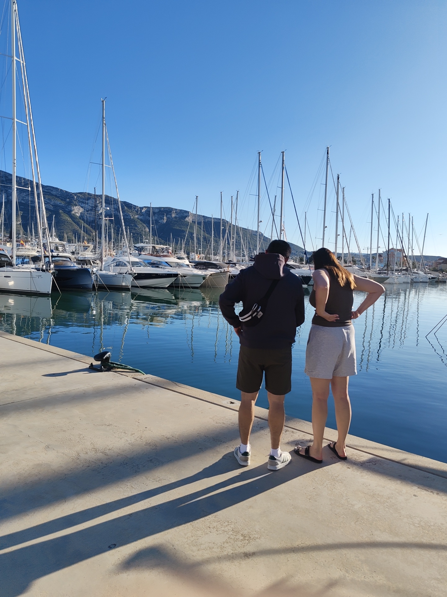 The image captures a bright, sunny day at a marina in Dénia, Spain, viewed from behind two people standing on a concrete dock. The man on the left wears dark shorts, a dark hooded jacket with a fanny pack slung over his shoulder, and sneakers. The woman on the right wears light grey shorts, a dark tank top, and black sandals. Both are looking out across the water towards a dense collection of moored boats.
The marina is bustling with numerous sailboats, their tall masts reaching towards a clear, deep blue sky, and a few motor yachts. The calm, blue water beautifully reflects the boats, masts, and rigging. In the background, rugged mountains with sparse green vegetation rise under the expansive blue sky.
Long shadows stretch from the individuals across the concrete dock in the foreground, indicating a low sun angle, characteristic of either morning or late afternoon. The scene conveys a peaceful atmosphere, with the clear skies and still water. Some boat registration numbers and names are faintly visible on the hulls of the vessels, such as "FLORINDA 2" and "7-P CP-2-206-17", but are not entirely distinct.