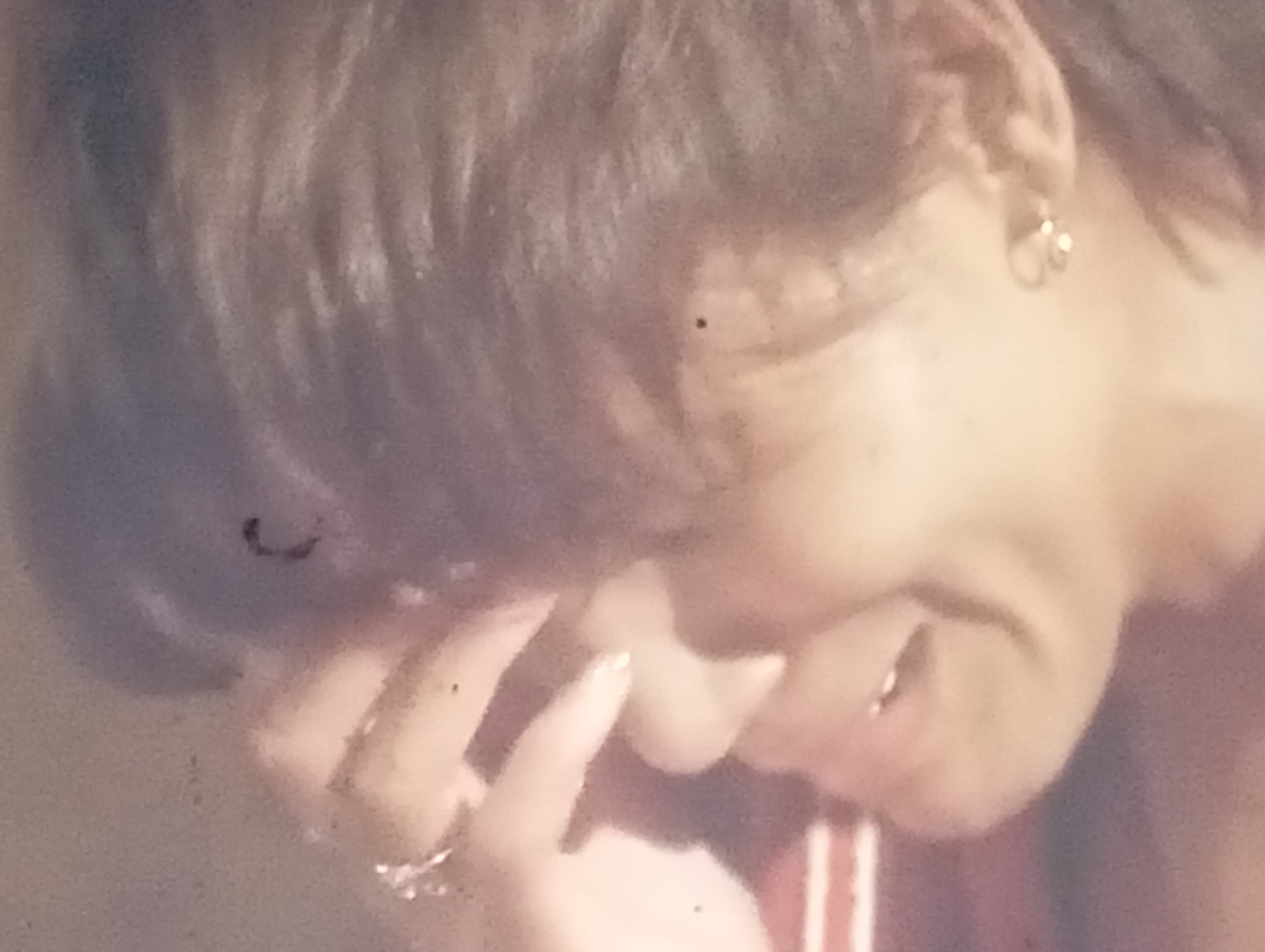 The image is a close-up, slightly blurry shot of a person's face, captured in what appears to be an indoor setting. The subject's hair is visible, styled in a way that suggests a modern haircut. They are wearing an earring on their visible ear, a small, circular hoop. The lighting is soft and warm, creating a slightly vintage feel to the image. The person's hand is brought up to their face, partially obscuring it, and their fingers are adorned with rings. The overall mood is intimate and perhaps pensive, as the subject's expression is partially hidden. The background is indistinct, with a gradient of colors that do not offer specific location cues. There is no visible text in the image. The context provided of Sofiivska Borshchahivka, Ukraine, does not directly inform the visual elements of the image itself.