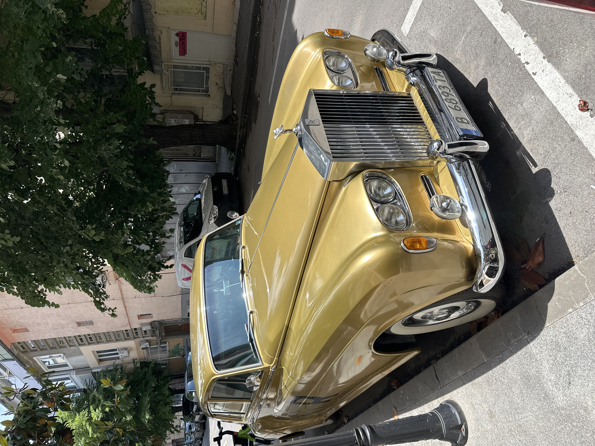 A gold Rolls-Royce, license plate B 6893 TA, is parked on a paved street in Varna, Bulgaria. The car is parked on the side of the street with a tree and buildings in the background. The chrome grille and hood ornament are visible. The street appears to be lined with other parked cars and trees. The image is taken during the day.