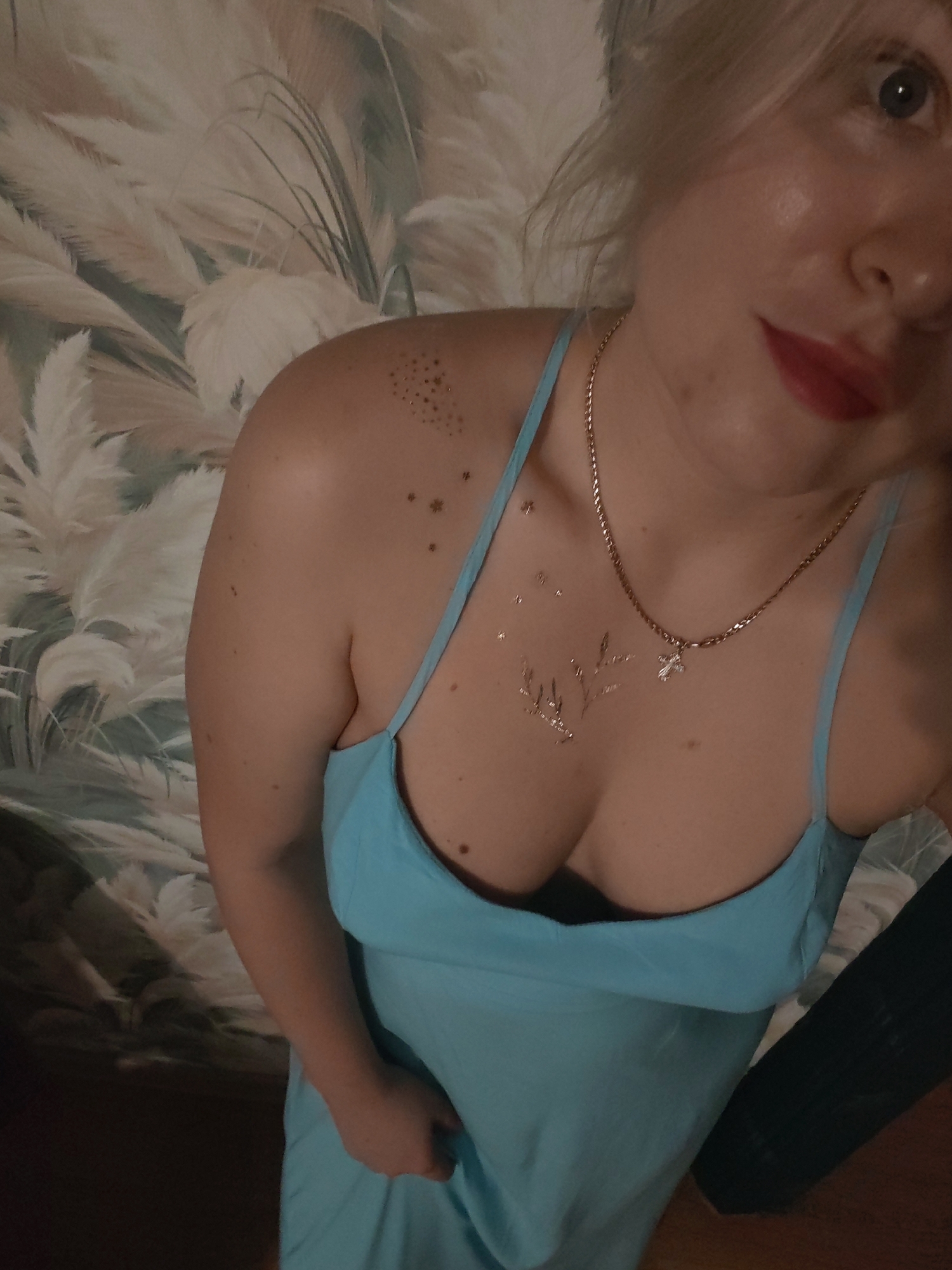 A woman with blonde hair and red lipstick is wearing a blue dress and a necklace with a cross pendant. She has glittery star and floral designs on her décolletage. The background is a wall with a floral pattern of beige and green reeds. The image is a close-up, taken from the chest up, and does not provide any information about the location in Izhevsk, Russia.