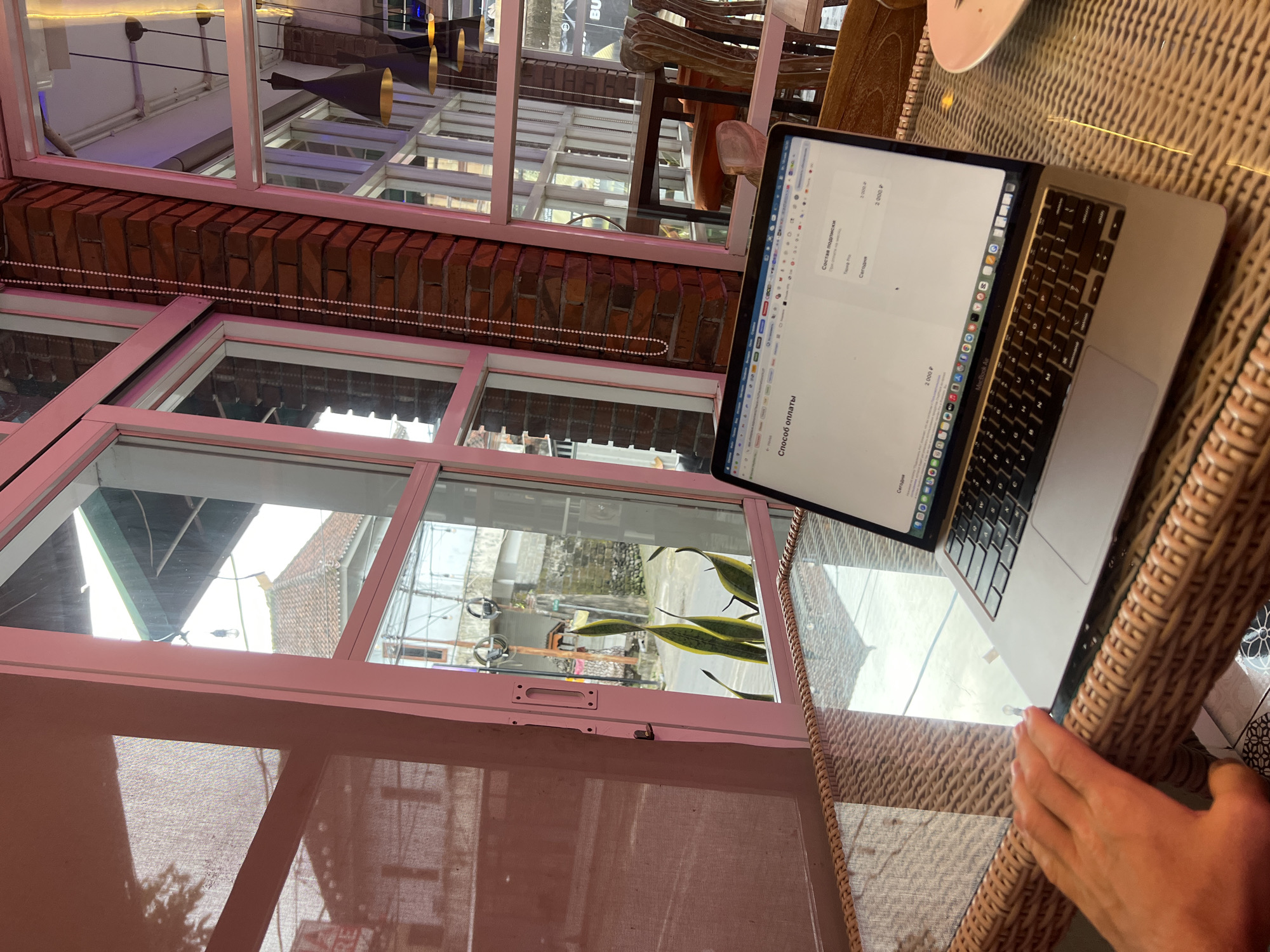 The image shows a person using a MacBook Air laptop while sitting at a table with a wicker surface, likely in a cafe or restaurant. The laptop screen displays text in a Cyrillic language. Outside the pink-framed window is a view of Sukawati, Indonesia, featuring buildings and greenery. The window's architecture includes a red brick section. The environment suggests an indoor-outdoor setting.