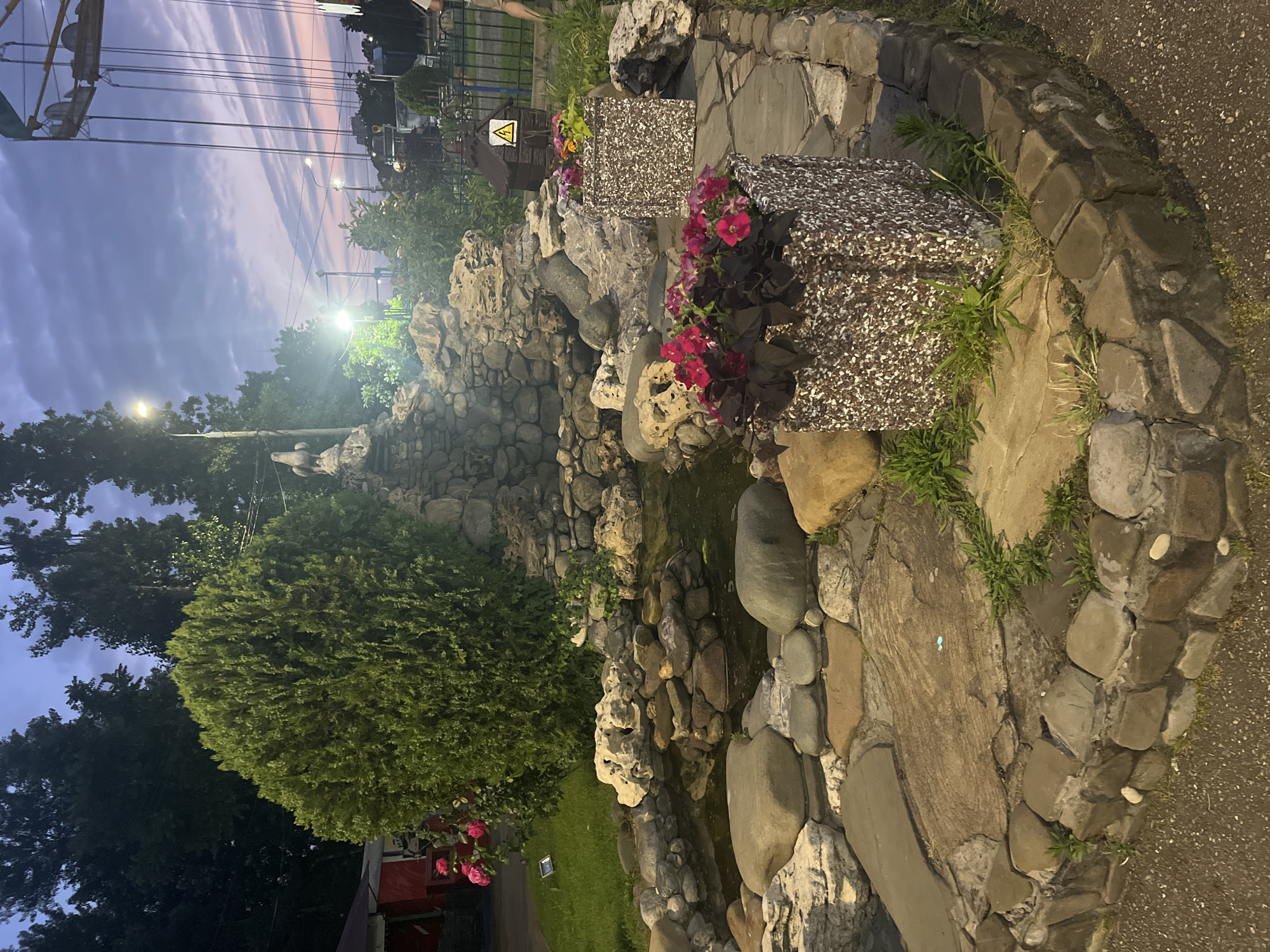 The image depicts an outdoor scene at dusk, possibly in a park or amusement area. In the foreground, there is a decorative rock garden with a small pond. Two square planters filled with pink and red flowers are placed on a stone pathway. Behind the rock garden, a large, manicured evergreen bush is visible, with pink roses growing nearby. In the background, a swing ride from an amusement park can be seen against a twilight sky with streaks of pink and purple. A street lamp illuminates the scene. There is a small, dark-colored structure with a yellow warning sign depicting a lightning bolt, and a statue of a bird atop a rock formation. The ground is a mix of gravel and stone paving. The overall atmosphere suggests a recreational setting at the end of the day. The information about the location in the city of Krasnodar, Russia, is not visually derivable from the image itself.