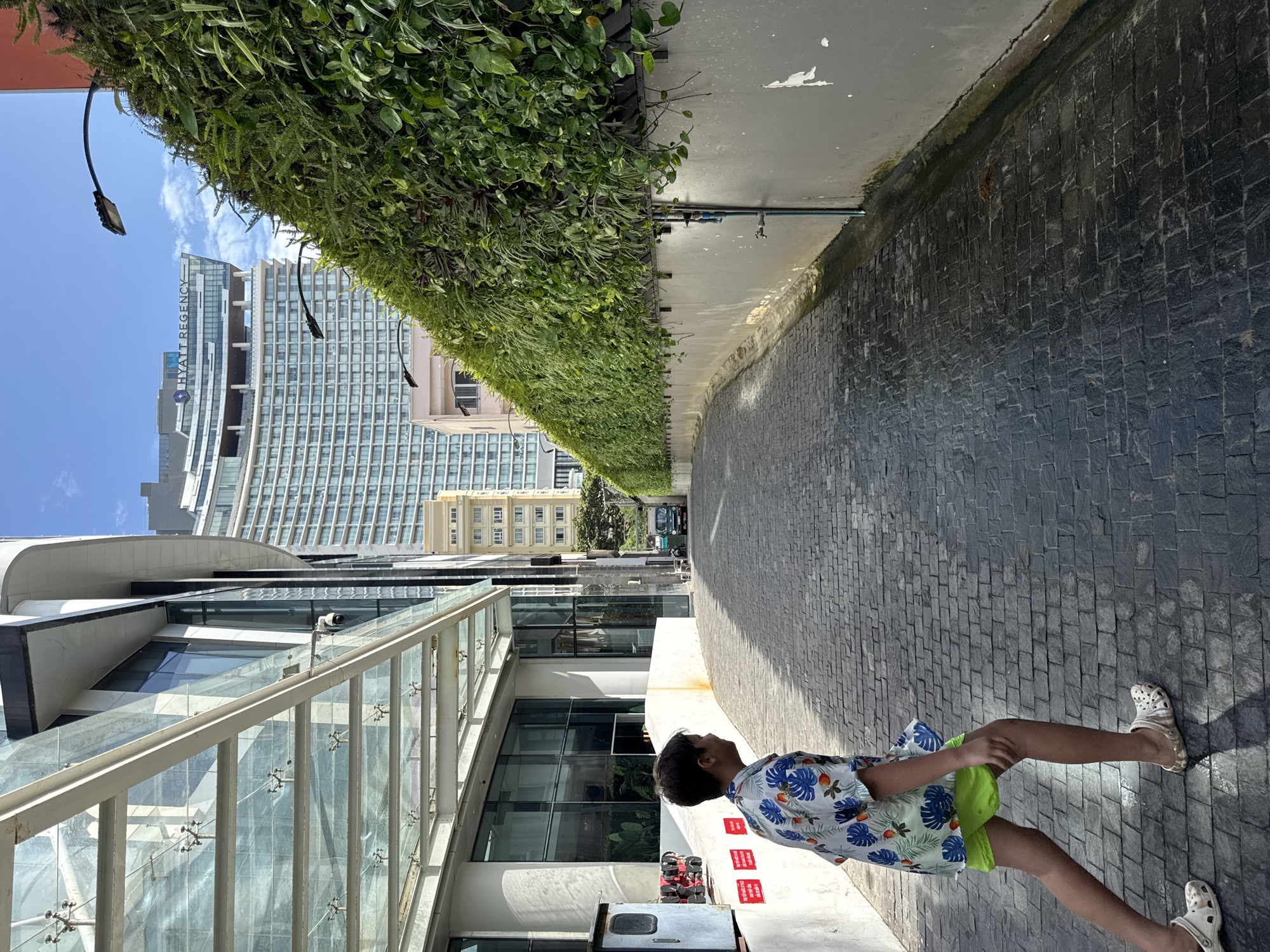 The image depicts a street scene in Nha Trang, Vietnam, with a child walking on a cobblestone pathway. The child, wearing a colorful short-sleeved shirt and bright green shorts, is facing away from the camera and is in motion. To the left of the pathway is a modern building with a glass canopy supported by white beams. On the right side of the pathway, a tall, grey wall is covered with a dense vertical garden of green plants. In the background, a large, contemporary hotel building with a curved facade, identified by a "HYATT REGENCY" sign, dominates the skyline. A few streetlights are visible along the top of the green wall, and a dark-colored van is parked further down the street on the right. The sky is blue with scattered clouds.