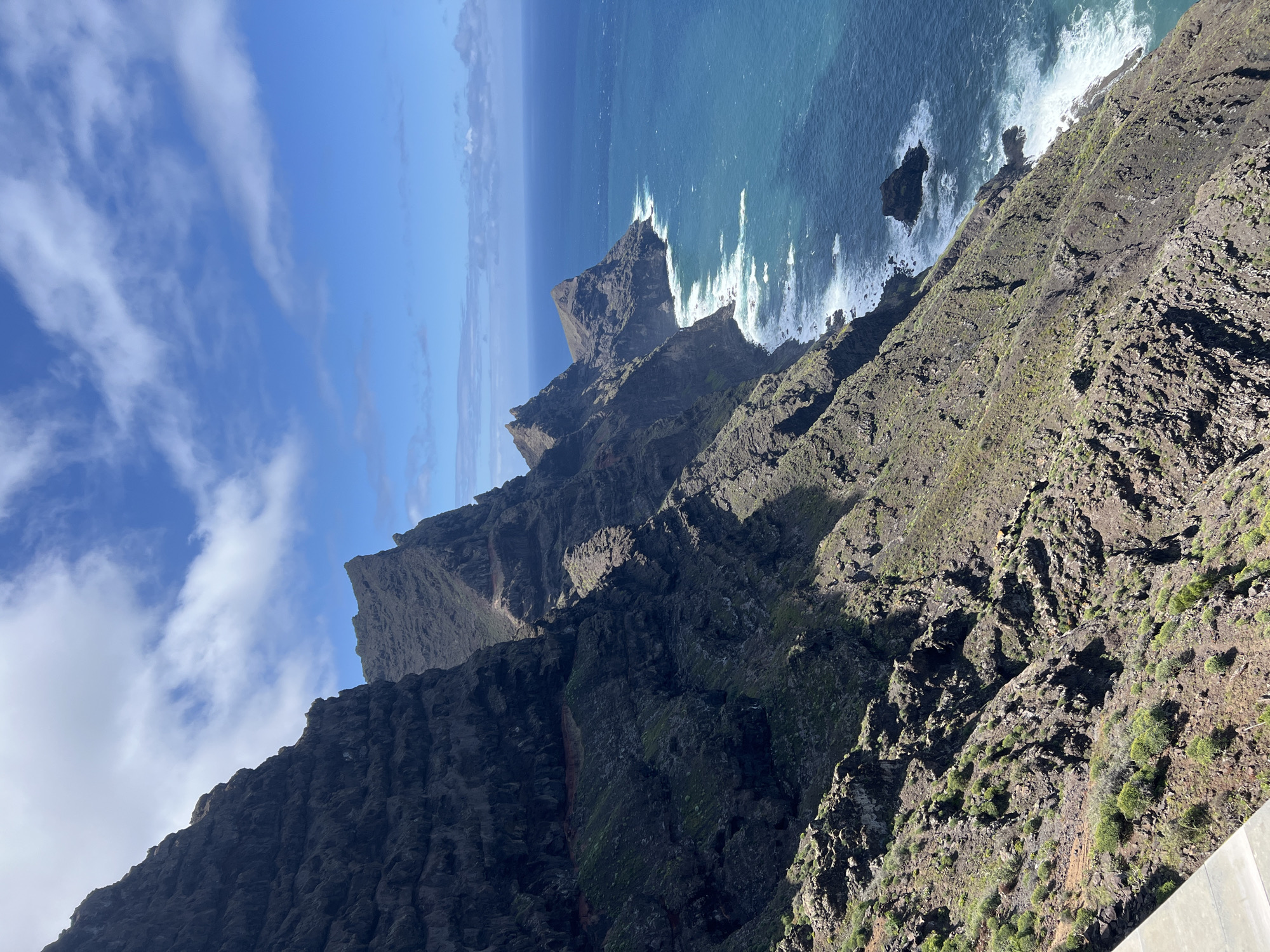 A high-angle, panoramic shot depicts a rugged coastline in Tirma, Spain. Jagged, dark grey and green cliffs plunge down to the turquoise ocean, where waves crash against the rocks. The sky is blue with scattered white clouds.