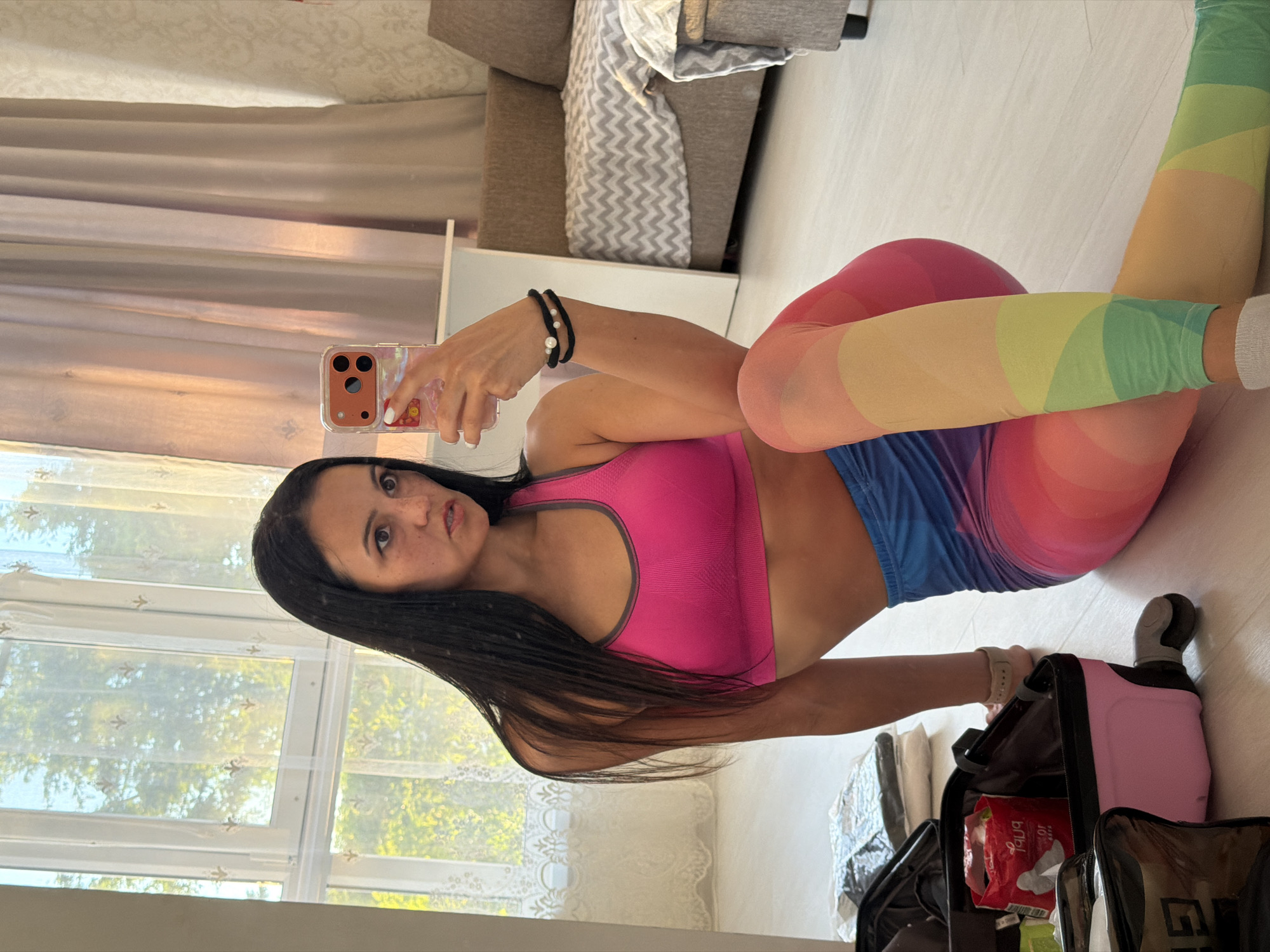 In this media file, a young woman is captured in a selfie, seemingly at home. She is wearing a bright pink sports bra and rainbow-patterned leggings, with blue shorts over them. Her dark hair is long and flows down her back. She is holding a smartphone in her hand, and her expression is direct, looking at the camera. The background shows a living room setting with a sofa and curtains, hinting at daytime with natural light filtering through a window. The woman has a casual and possibly playful demeanor.
The scene appears to be indoors, likely a residential setting. The presence of a sofa and curtains suggests a living area. The bright lighting implies it is daytime. There are no overt indications of specific weather conditions, but the natural light suggests it is not nighttime or heavily overcast. The woman is posing for a selfie, indicating a personal moment captured for social media or personal record.
Notable details include the woman's attire, the rainbow pattern of her leggings, and the pink sports bra. The smartphone she is holding has a distinct camera setup. The background is slightly blurred but provides context of a domestic environment.
The visible text is limited to the brand name on the red package inside the bag, which appears to be "Always". The location context is Sochi, Russia.