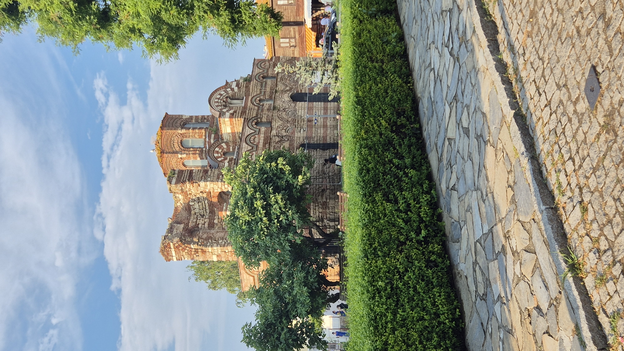 The image depicts the Church of St. Stephen in Nesebar, Bulgaria, a UNESCO World Heritage site. The ancient, partially ruined brick structure, characterized by its arched windows and intricate brickwork, dominates the upper half of the frame. The building is surrounded by greenery, with lush trees and a well-maintained hedge in the foreground. A cobblestone path leads away from the viewer, bordered by a stone walkway. Several individuals are visible in the background, some seated on benches and others walking, suggesting a public space. The sky above is blue with scattered clouds. The overall scene conveys a sense of historical significance and tranquil outdoor atmosphere.