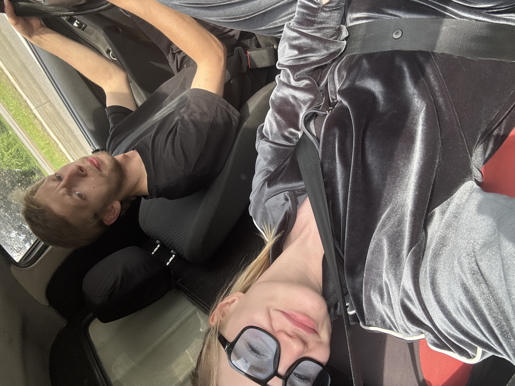 In this overhead, close-up shot, two individuals are seated inside a vehicle. The person on the left, facing the camera, wears a grey velour tracksuit and black-rimmed glasses. Their eyes are looking slightly upward and to the right, with a subtle smile on their face. A black seatbelt is visible across their chest. To the right, a person with light brown hair wears a black t-shirt. Their gaze is directed towards the camera with a slight frown and their hands are positioned on what appears to be a steering wheel. The background shows car interior elements, including a headrest and a seat, with a glimpse of greenery and a road visible through the side window.