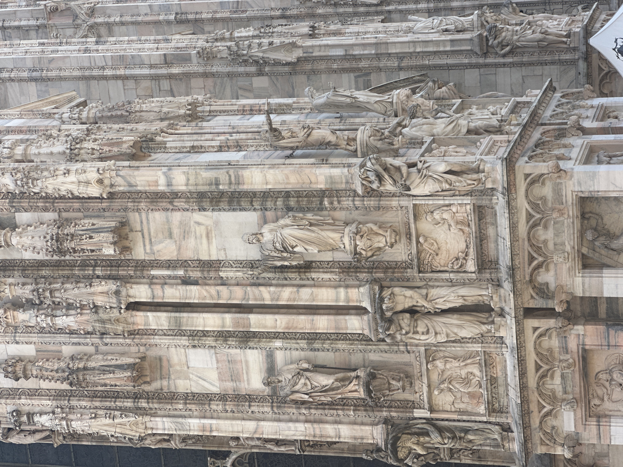 This is a detailed, close-up, exterior, eye-level, full-shot photograph of the side facade of the Duomo di Milano (Milan Cathedral) in Milan, Italy. The cathedral is constructed of a pale marble with pink and grey veining. The facade is heavily adorned with gothic architectural elements, including intricate carvings, niches with statues, pinnacles, and decorative mouldings.
There are numerous statues of various sizes and compositions depicted. Some are full-length figures of men in flowing robes, standing on pedestals. One statue on the left appears to be of a standing figure in a robe, slightly obscured by the edge of the frame. Further to the right, there is a statue of a man holding a staff. To his right, another man in a robe is pointing with his right arm. Another statue further right shows a man with a beard, looking downwards. There are also bas-relief carvings depicting scenes with multiple figures, some appearing to be religious in nature.
The architecture exhibits a strong vertical emphasis with repetitive decorative patterns and columns. The lighting is diffused, suggesting an overcast or indirectly lit environment. The overall impression is one of grandeur and detailed craftsmanship associated with Gothic architecture. There are no visible people actively engaged in any actions. The scene is static and architectural.
