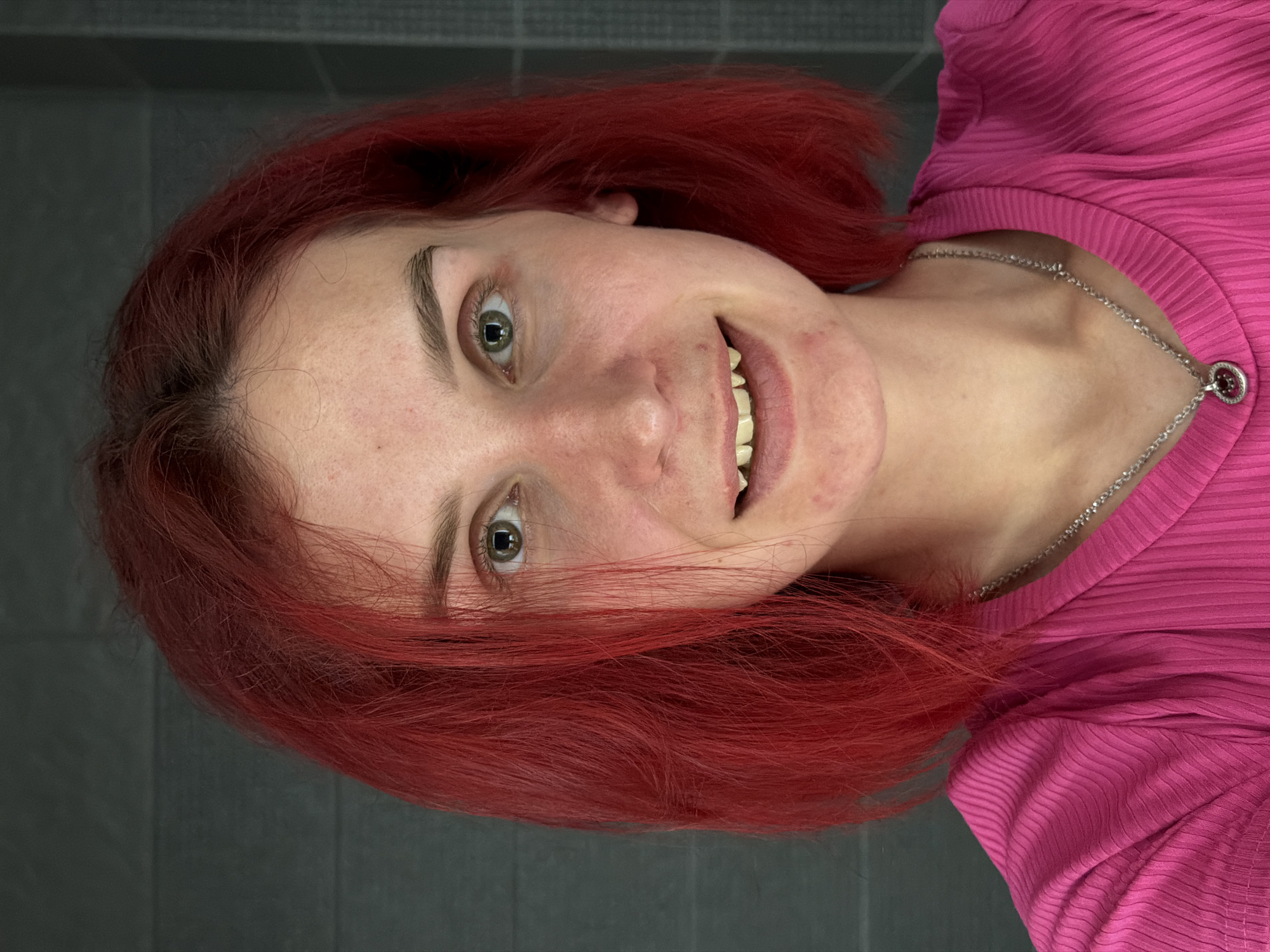 A young woman with red hair and fair skin is facing the camera, slightly rotated. She is wearing a pink ribbed shirt and a silver necklace. Her eyes are light-colored, and she is smiling, revealing her teeth. The background is a grey wall with a subtle textured pattern. There is no clear evidence to determine if this photo was taken in the city of Chaikovsky, Russia.