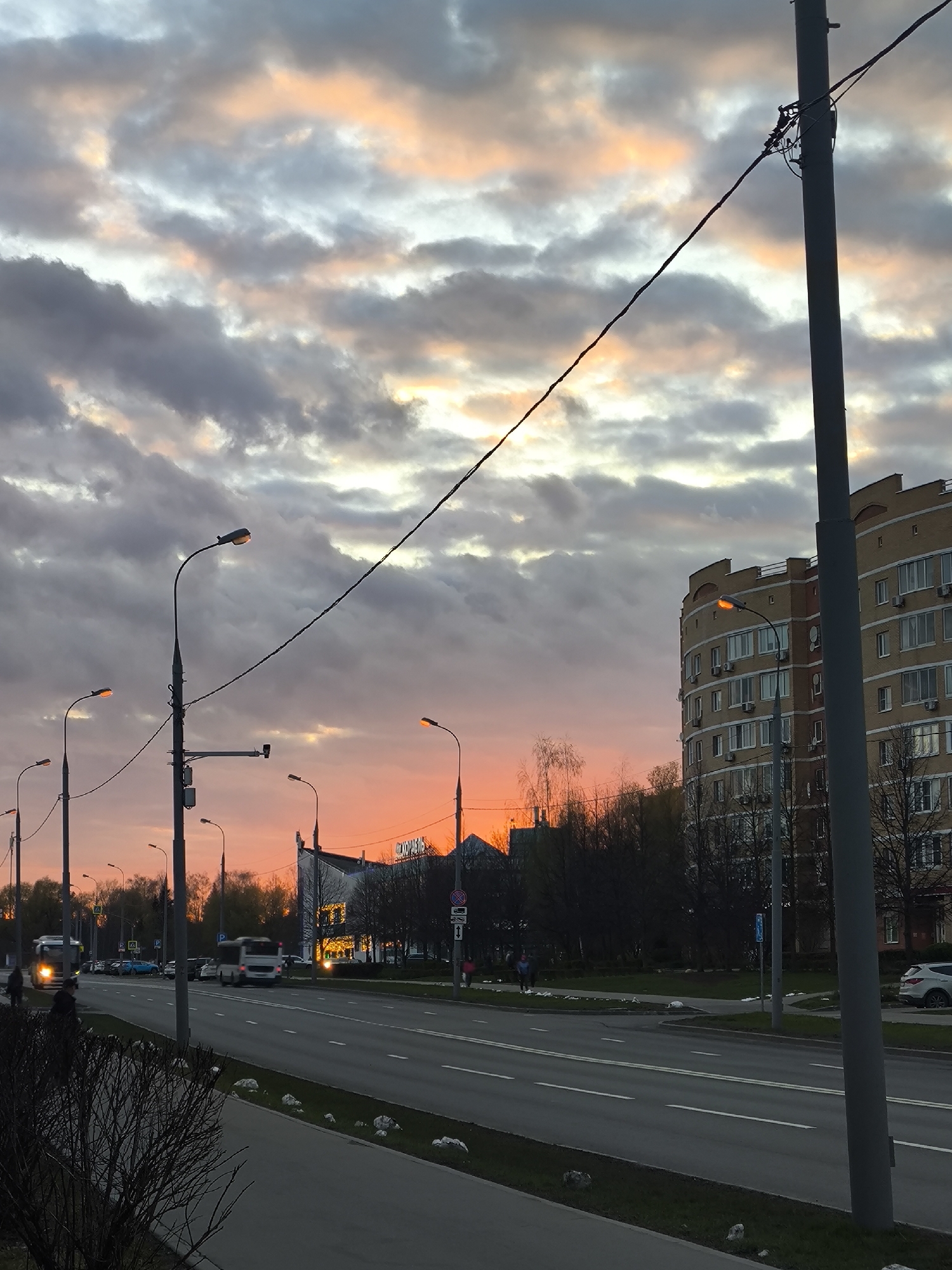 This image captures an urban street scene in Moscow, Russia, during twilight hours, likely sunset, indicated by the vibrant orange and pink hues painting the underside of the heavy grey clouds. Numerous streetlights are illuminated, casting a warm glow on the surroundings.
A multi-lane asphalt road with clear white markings, including a double solid yellow line in the center, dominates the mid-ground. Several vehicles, including cars and a white bus, are in motion or parked along the road. On the right, a prominent multi-story residential building features a curved, light beige brick facade with many windows and visible air conditioning units. Further down the road on the left, a darker building with brightly lit windows stands out, displaying Cyrillic text "СКОРОСТЬ" (SKOROST', meaning "SPEED") on its exterior.
The scene is lined with bare trees and bushes, suggesting either late autumn or early spring. Tall utility poles with overhead wires crisscross the sky, adding to the urban infrastructure. Concrete sidewalks are visible on both sides of the road, with grass verges. Several people can be seen walking on the sidewalks, their figures somewhat blurred by motion. The overall impression is one of an active city transitioning from day to night under a dramatically colored sky.
