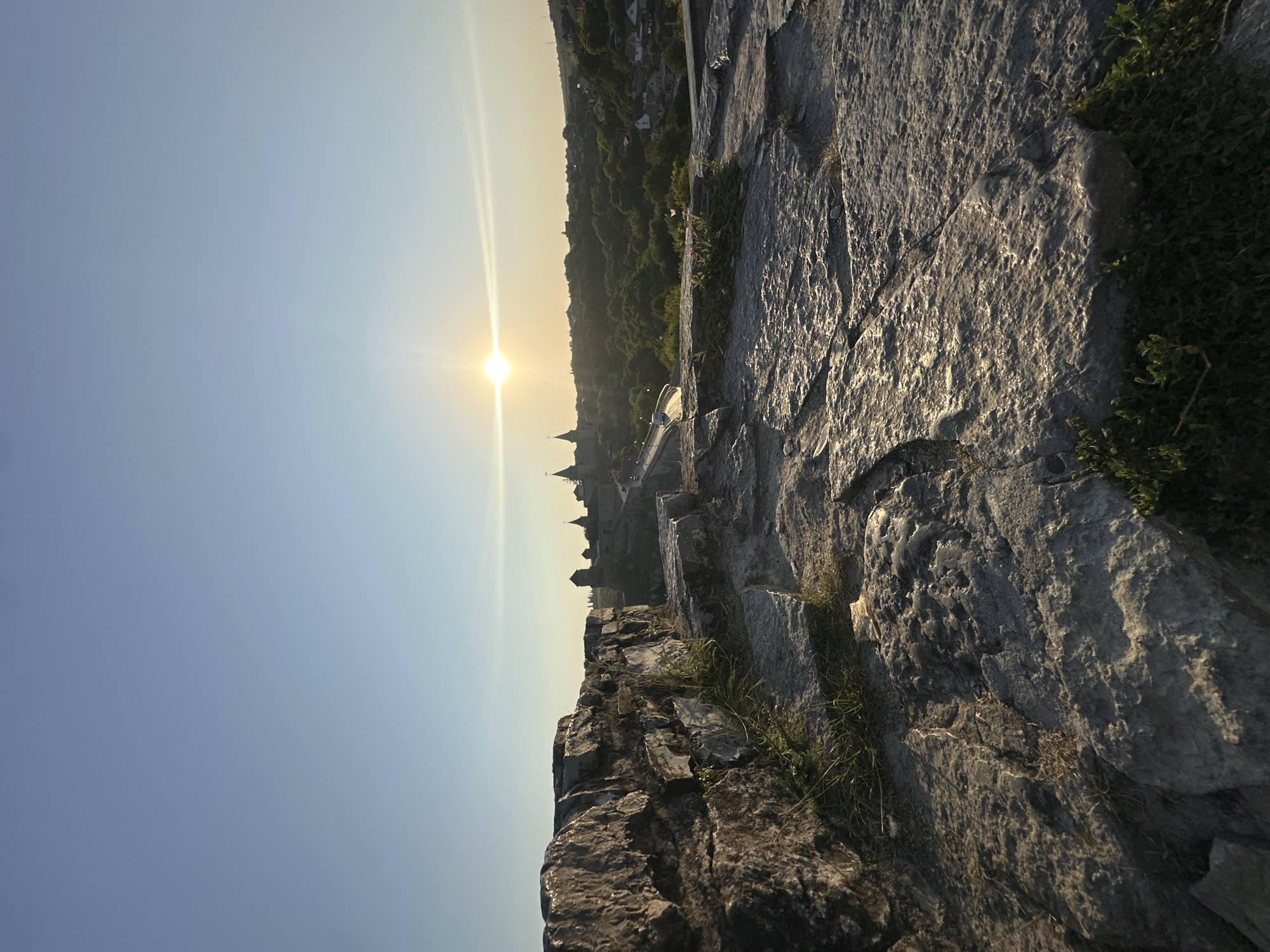 The image captures a scenic view of Kamianets-Podilskyi, Ukraine, at sunset. The foreground is dominated by rough, textured stone formations, possibly part of the ancient fortress walls, with sparse patches of green grass growing between the rocks. In the mid-ground, a bridge extends across a ravine, leading towards the iconic Kamianets-Podilskyi Castle, its spires silhouetted against the warm, golden glow of the setting sun. The castle complex appears ancient and majestic, a testament to the city's rich history. Beyond the castle, a lush green landscape unfolds, dotted with trees and scattered buildings that suggest the presence of a town or village nestled in the valley. The sky above is a clear, pale blue, transitioning into a lighter hue near the horizon where the sun is setting, casting long, dramatic light rays. The overall atmosphere is serene and picturesque, highlighting the architectural marvel of the castle and its natural surroundings bathed in the soft light of dusk. There are no people visible in the scene.