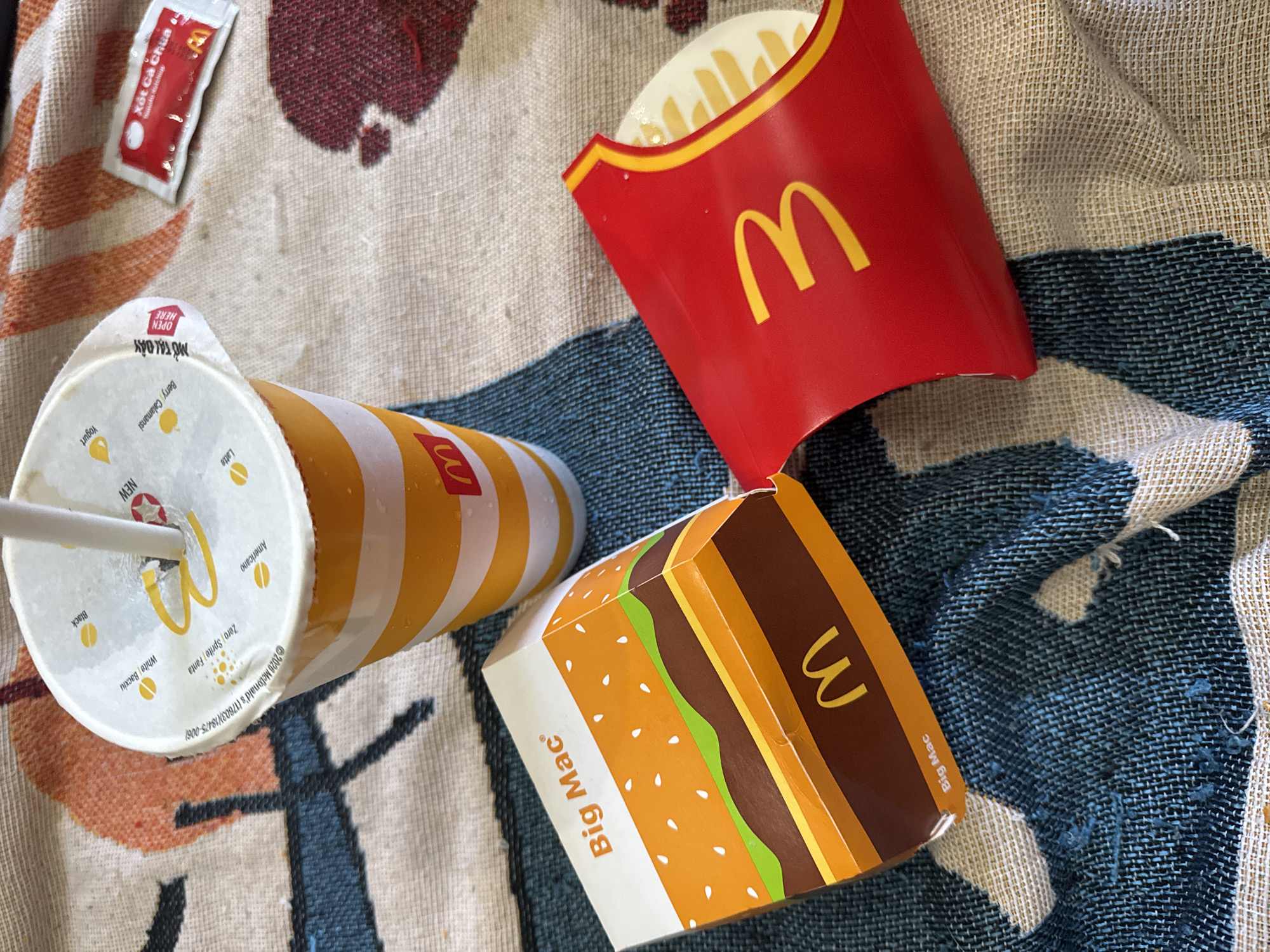 A McDonald's Big Mac box, a red French fry container with fries, and a McDonald's branded cup with a straw are arranged on a textured, patterned blanket. A packet of ketchup is also visible on the blanket. The image is taken from a high angle, with the items positioned in the center. The background is blurred, suggesting the focus is on the food items. There are no people visible in the image.