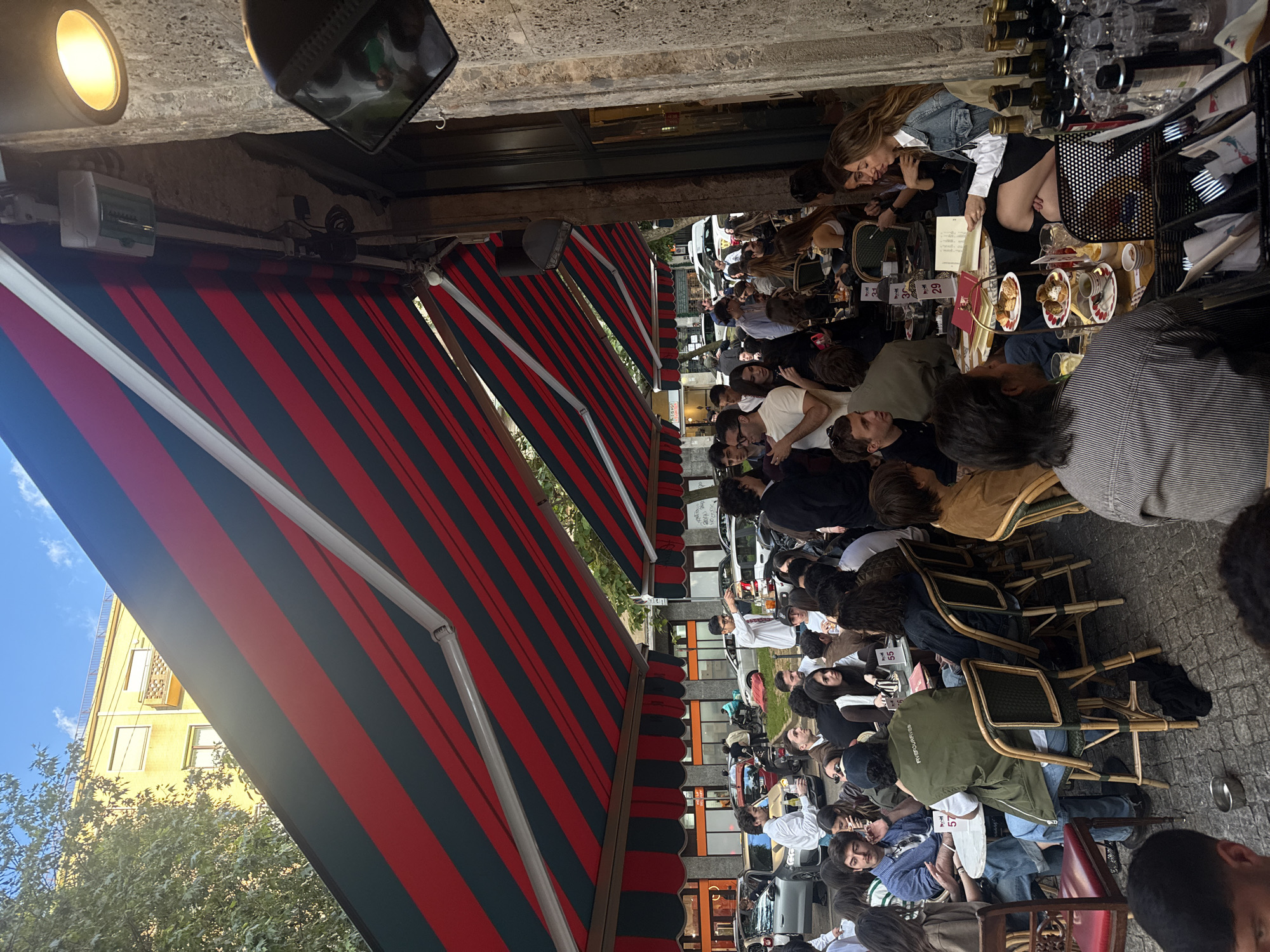 A bustling outdoor cafe scene unfolds beneath a red and black striped awning in Milan, Italy. Patrons are seated at closely arranged tables, enjoying food and conversation. Many individuals are visible, exhibiting a range of ages and attire. The cafe's furnishings include wicker chairs. The street is visible beyond the tables, lined with parked vehicles and buildings. Wine bottles and glassware are positioned near the tables. Overhead, a light fixture is mounted to the building.
