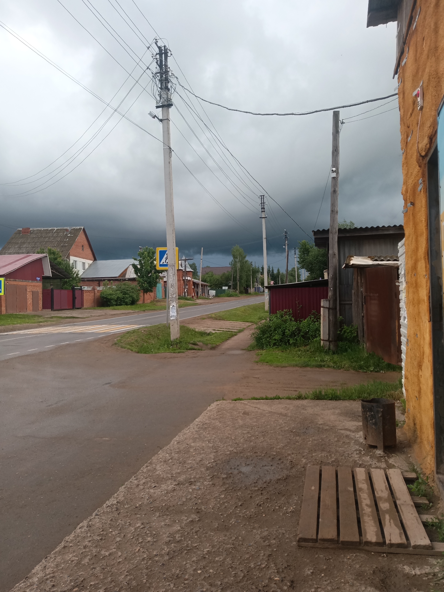 This image captures a quiet, somewhat gloomy street scene in a rural or semi-rural area, identified as Фоки, Russia. The dominant feature is the heavily overcast sky, filled with dark, ominous storm clouds that suggest an impending or ongoing storm, casting a dim light over the entire landscape.
In the foreground, a paved road with a prominent pedestrian zebra crossing runs across the frame. A dirt path or shoulder extends along the roadside. To the far right, part of a building with a textured, orange-yellow wall is visible, with a wooden pallet-like structure and a metal bin on the ground nearby.
Multiple utility poles, heavily laden with intricate power and communication lines, crisscross the sky. One large, central utility pole stands prominently on the left side of the road, featuring a complex network of wires and equipment. A smaller wooden pole closer to the right also holds lines and a security camera.
Beyond the road, various residential structures are visible, including brick houses with differing roof styles (some red-tiled, others grey), and what appear to be sheds or garages. A red metal gate and brick fences mark property boundaries. Green trees and bushes are interspersed throughout the scene. Two yellow square road signs with blue pedestrian crossing icons are strategically placed to indicate the zebra crossing. No people are visible, and the scene conveys a sense of stillness, perhaps in anticipation of the storm's arrival. The road surface appears slightly damp under the heavy cloud cover.