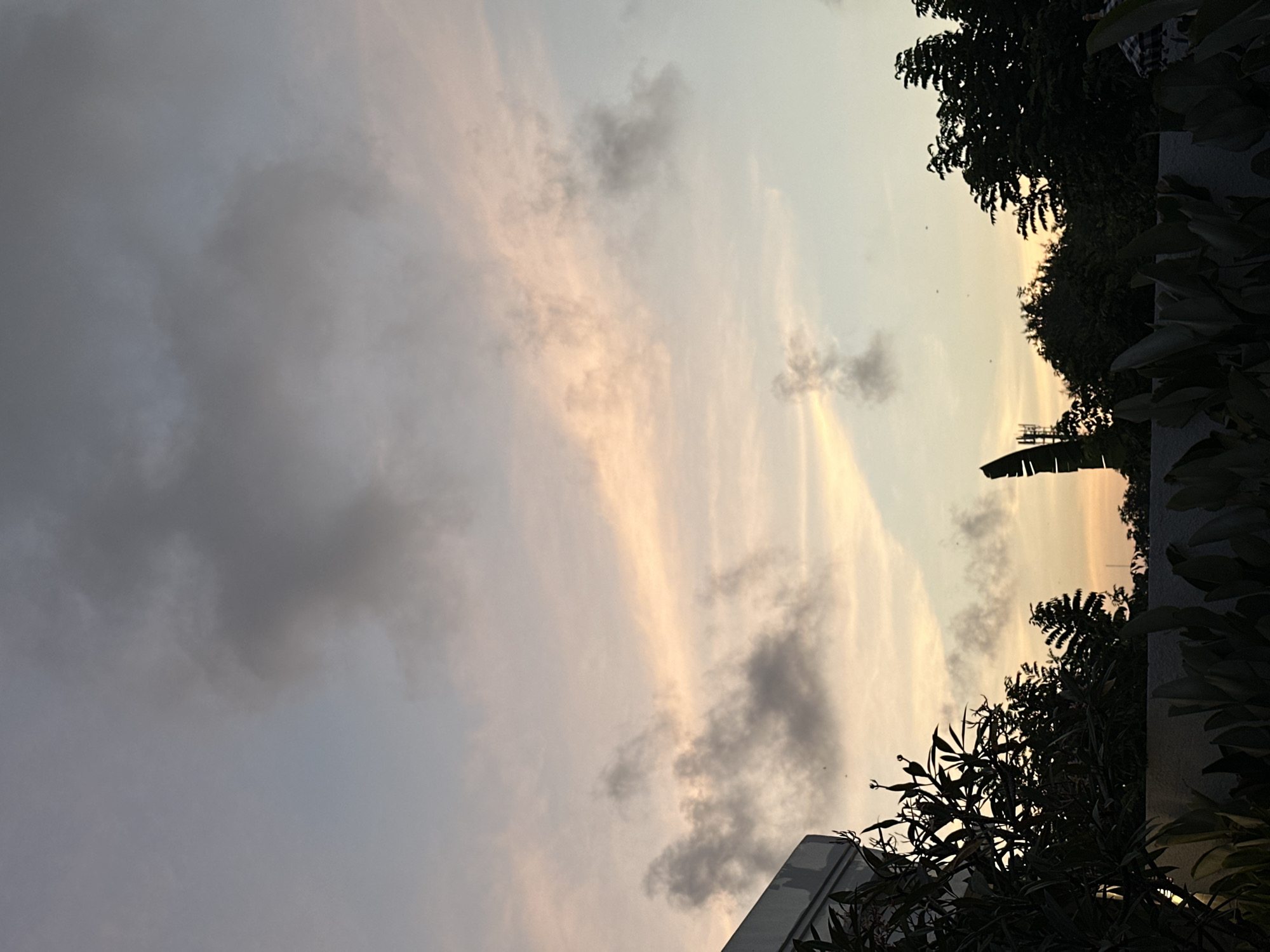 The image depicts a sky filled with clouds at sunset, with hues of pink, orange, and blue. Silhouetted trees and foliage form the foreground, with a building visible on the left. The setting appears to be an outdoor area, possibly a rooftop or balcony, in Kuta Selatan, Indonesia. There are no people or specific actions discernible in the scene. The overall mood is serene and tranquil, typical of a tropical evening.