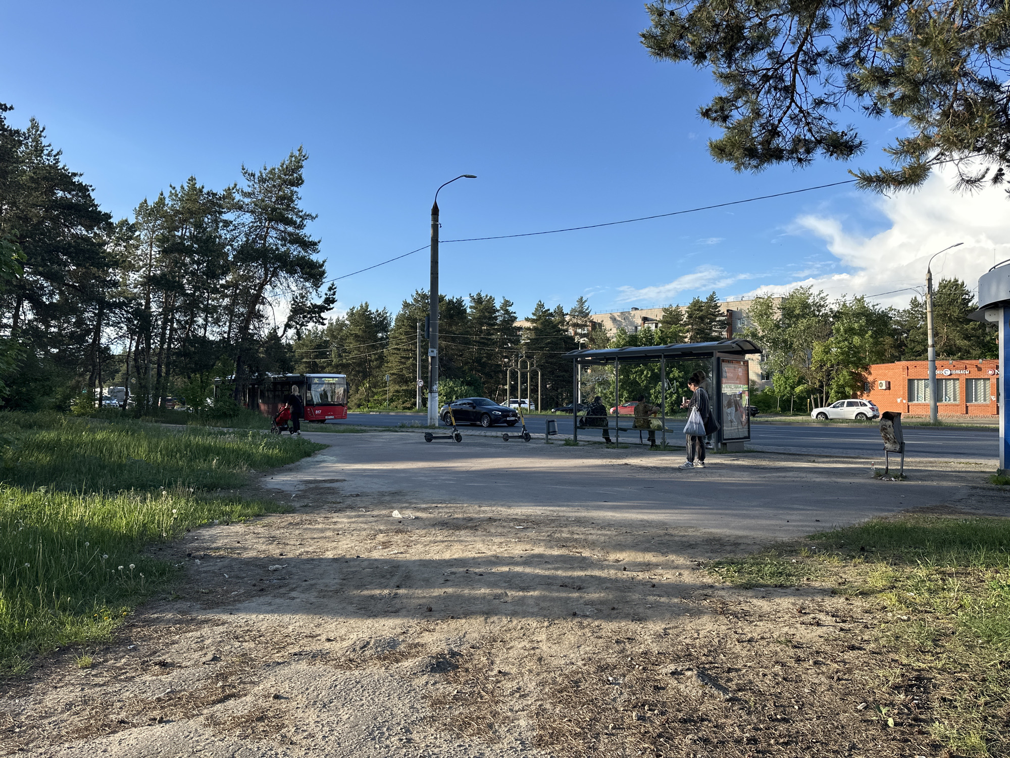 The scene depicts a bus stop in what appears to be a suburban or peri-urban area of Vladimir, Russia. A red city bus is visible on the left side of the road, partially obscured by trees. Several individuals are present at the bus stop, some seated under the shelter and one person standing. Parked scooters are beside the shelter. A black car is on the road. A light pole stands tall behind the bus stop. Across the road, there is a red brick building, possibly a store, with signage in Russian. The ground in the foreground is a mixture of dirt and sparse grass. The sky is blue with scattered clouds. The general atmosphere is that of an ordinary day at a public transport stop.