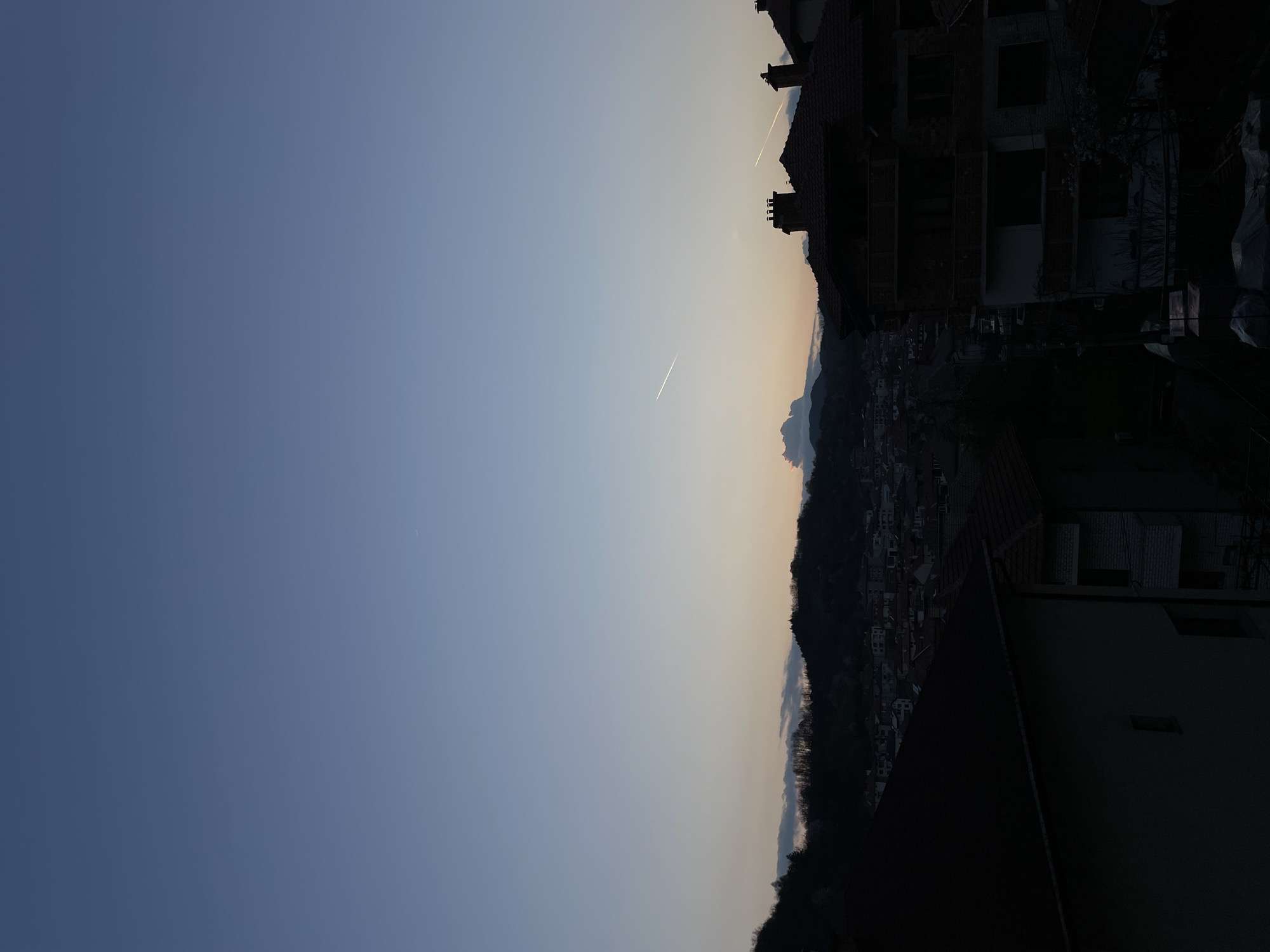 The image shows an aerial view of Zlatograd, Bulgaria. The sky is a gradient of blue to orange, indicating sunrise or sunset. There are two contrails visible in the sky. The skyline is composed of mountains and buildings, with some under construction. The scene appears to be captured from a high vantage point, such as a rooftop. Details of the city below are obscured by shadows.