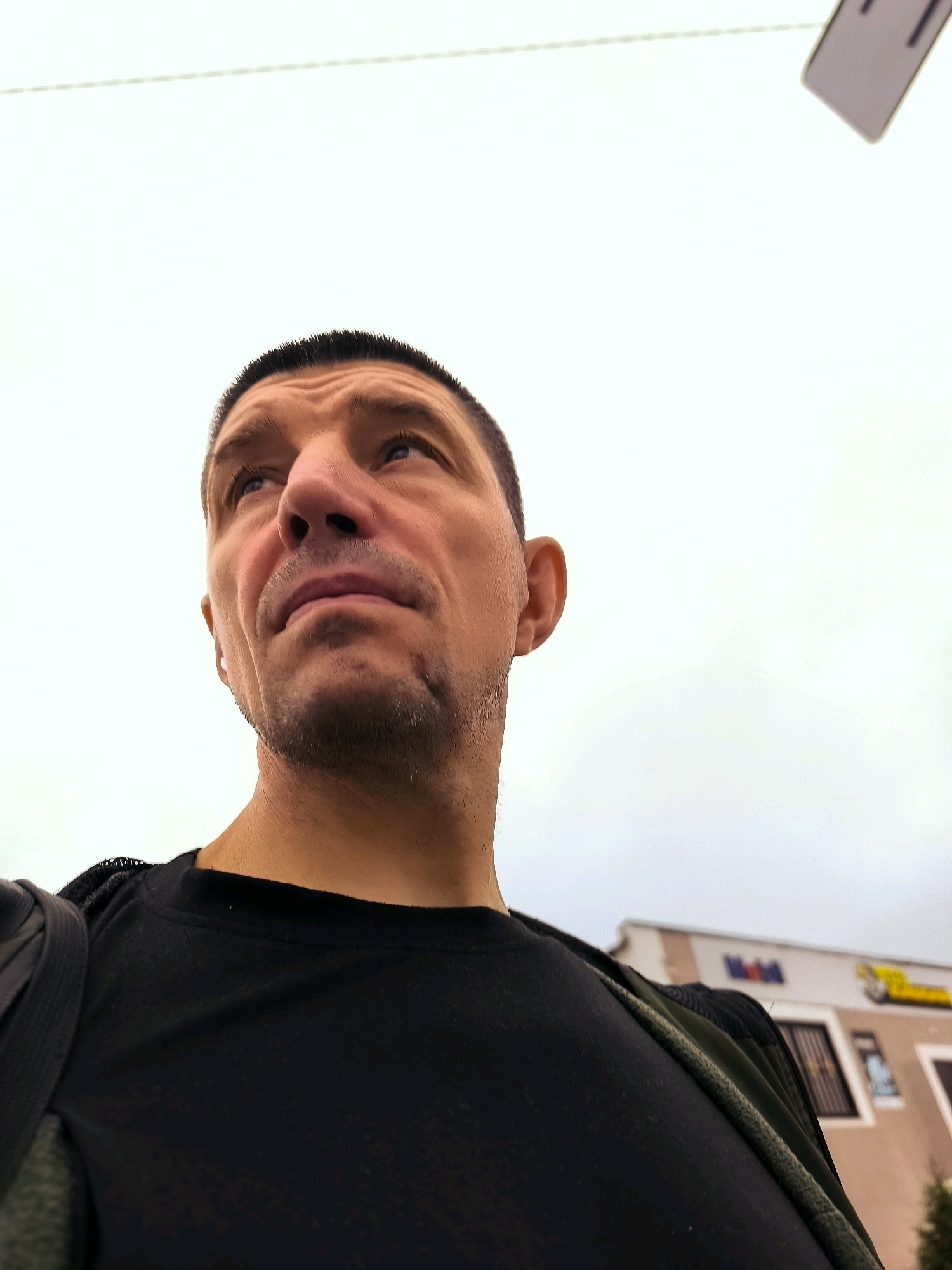 A close-up, low-angle shot captures a man's face from the chest up, looking upwards and to the left. The man has short, dark hair, a prominent nose, and is wearing a black t-shirt and a backpack strap. A blurred building with a "Mobil" sign is visible in the background, suggesting an urban outdoor setting. The sky is overcast. The image does not depict any specific location within the Qarağandı city administration, Kazakhstan.
