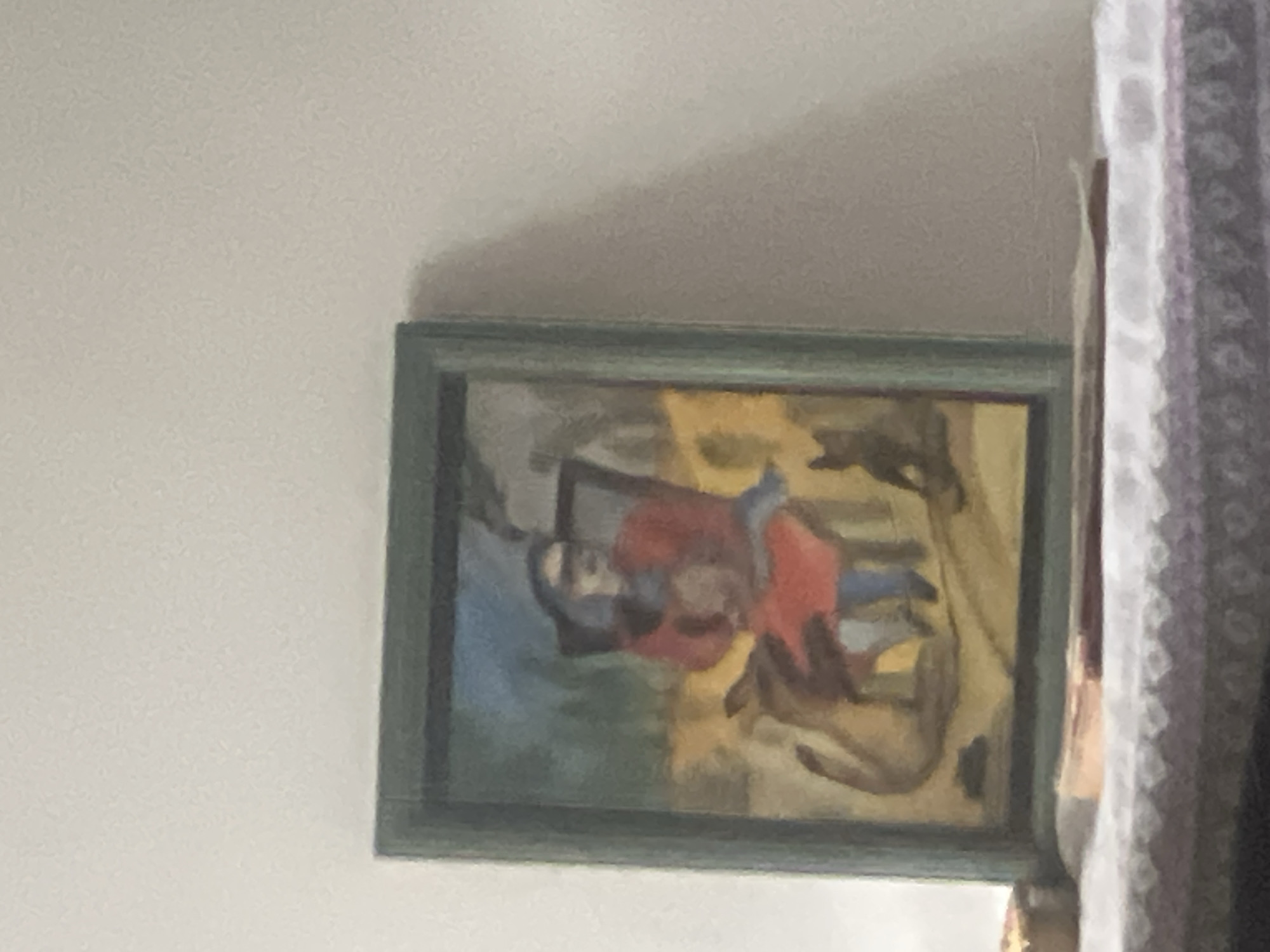 The image displays a framed painting resting on a white, patterned tablecloth, positioned against a light-colored, textured wall in Casablanca, Morocco. The painting, rendered in a folk art style, depicts a central figure seated in a chair, wearing a red top and blue lower garment, with darkened or obscured facial features. To the left of the seated figure, a large, light brown quadruped with a long tail stands, while a smaller, dark quadruped sits to the right. The background of the painting features a blue sky or upper section above a yellow-brown ground, with a dark, vertical element resembling smoke or a distant structure rising from the upper right. The painting is encased in a dark green frame. On the tablecloth directly in front of the painting, two stacked, flat objects, consistent with books or papers, are visible. A small, light-colored, cylindrical object is partially visible on the tablecloth to the left of the frame. The scene is illuminated by soft, ambient light.