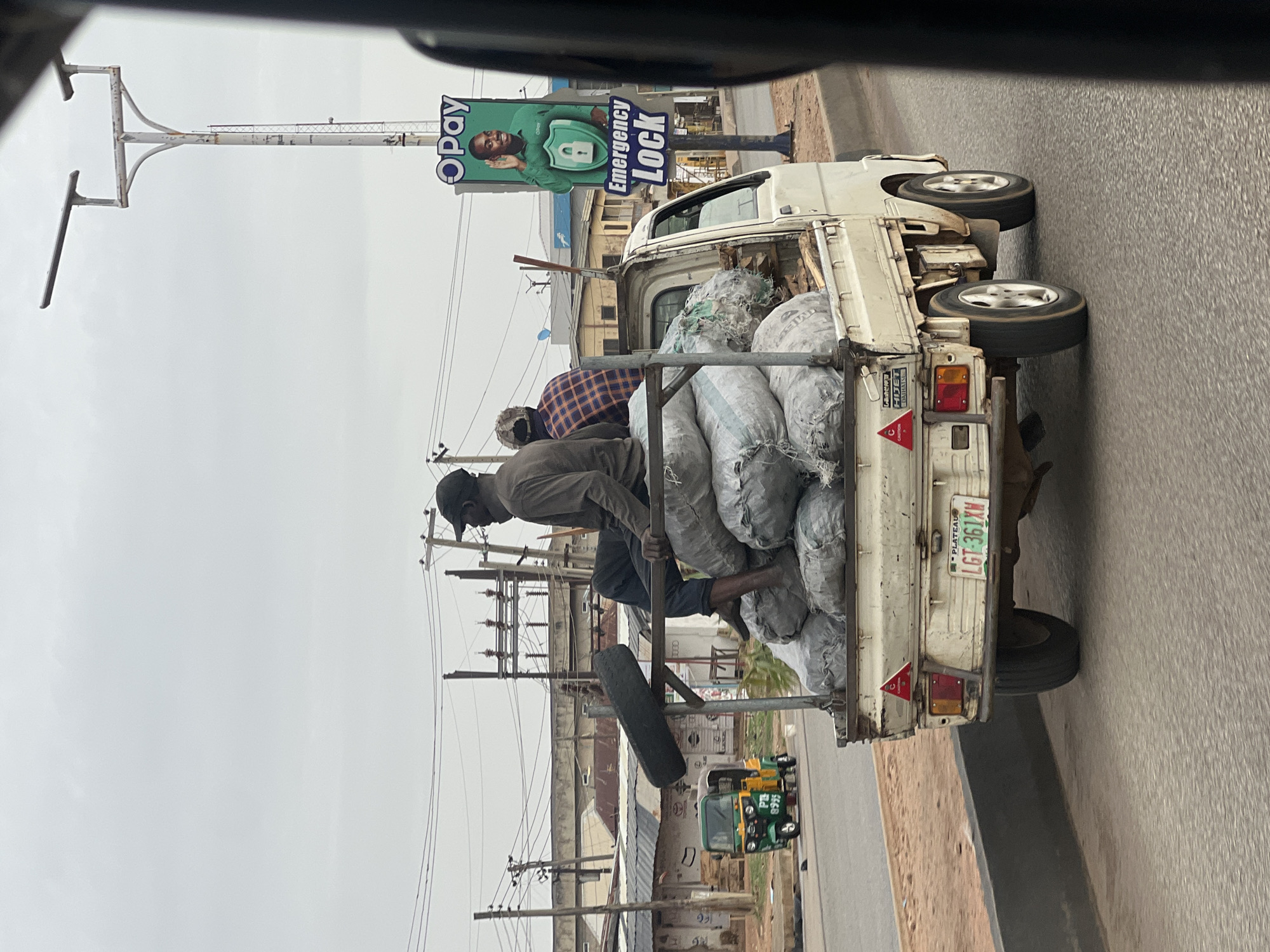 The image depicts a small, white flatbed truck overloaded with large sacks, presumably containing charcoal or similar goods. Two individuals are riding atop the load. The truck is traveling on a paved road. A billboard advertising "Opay" with the words "Emergency Lock" is visible in the background. Electrical power lines are present along the roadside. A three-wheeled vehicle is visible in the background. The license plate on the truck reads "LGT-361XM," and "Plateau" is written at the top of the plate. Based on the vehicle's license plate, the image was taken in the vicinity of Gura Top, Nigeria.