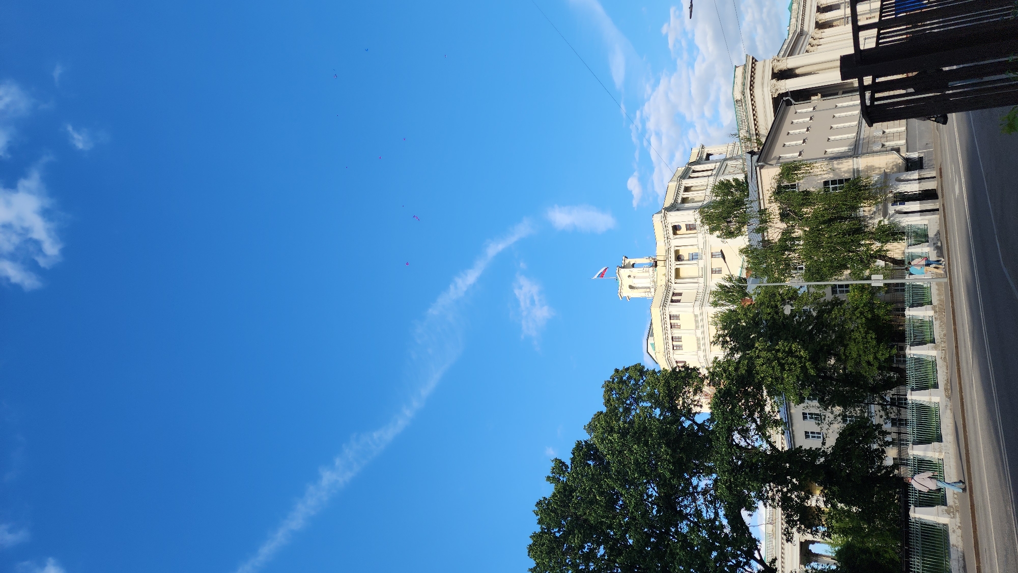 The image shows a portion of a building complex in Moscow, Russia, under a blue sky with scattered clouds and contrails. A Russian flag is visible on the roof of one of the buildings. Trees partially obscure the lower sections of the buildings, which are separated from the street by a wrought iron fence. Two pedestrians walk along the sidewalk adjacent to the road. The image is rotated approximately 90 degrees counter-clockwise. The vantage point is likely from an elevated position. Several small red objects are visible in the sky.