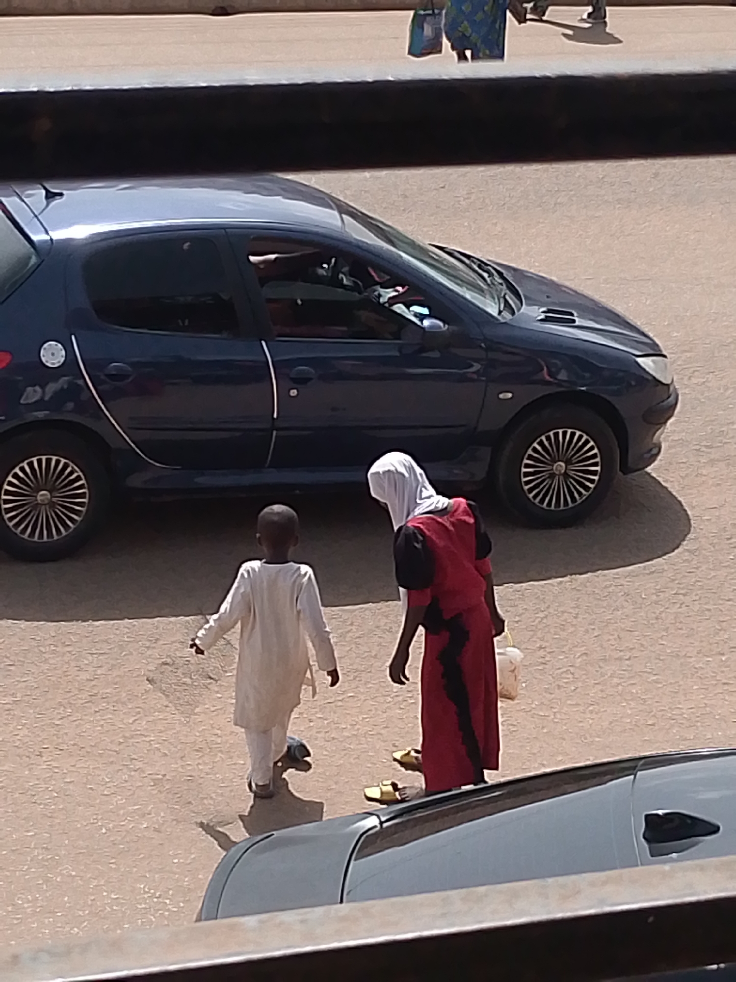This media file depicts a street scene in Bukuru, Nigeria. Two individuals, a young boy and an adult wearing a hijab and a red dress, are walking. The boy is clad in a white garment and appears to be walking away from the camera. The adult is wearing a white hijab and a long red dress with black accents, and is carrying a light-colored bag in their right hand. They are both on a paved surface, likely a street or walkway, with a dark blue car parked nearby. The car has its doors closed and appears to be stationary. In the background, the blurry shapes of other people and possibly buildings are visible, suggesting an urban or semi-urban environment. A portion of a gray vehicle is also visible in the foreground, partially obscuring the lower part of the scene. The lighting suggests it is daytime, with the sun casting shadows on the ground. No specific events are clearly taking place, but the individuals appear to be walking. There is no visible text in the image.